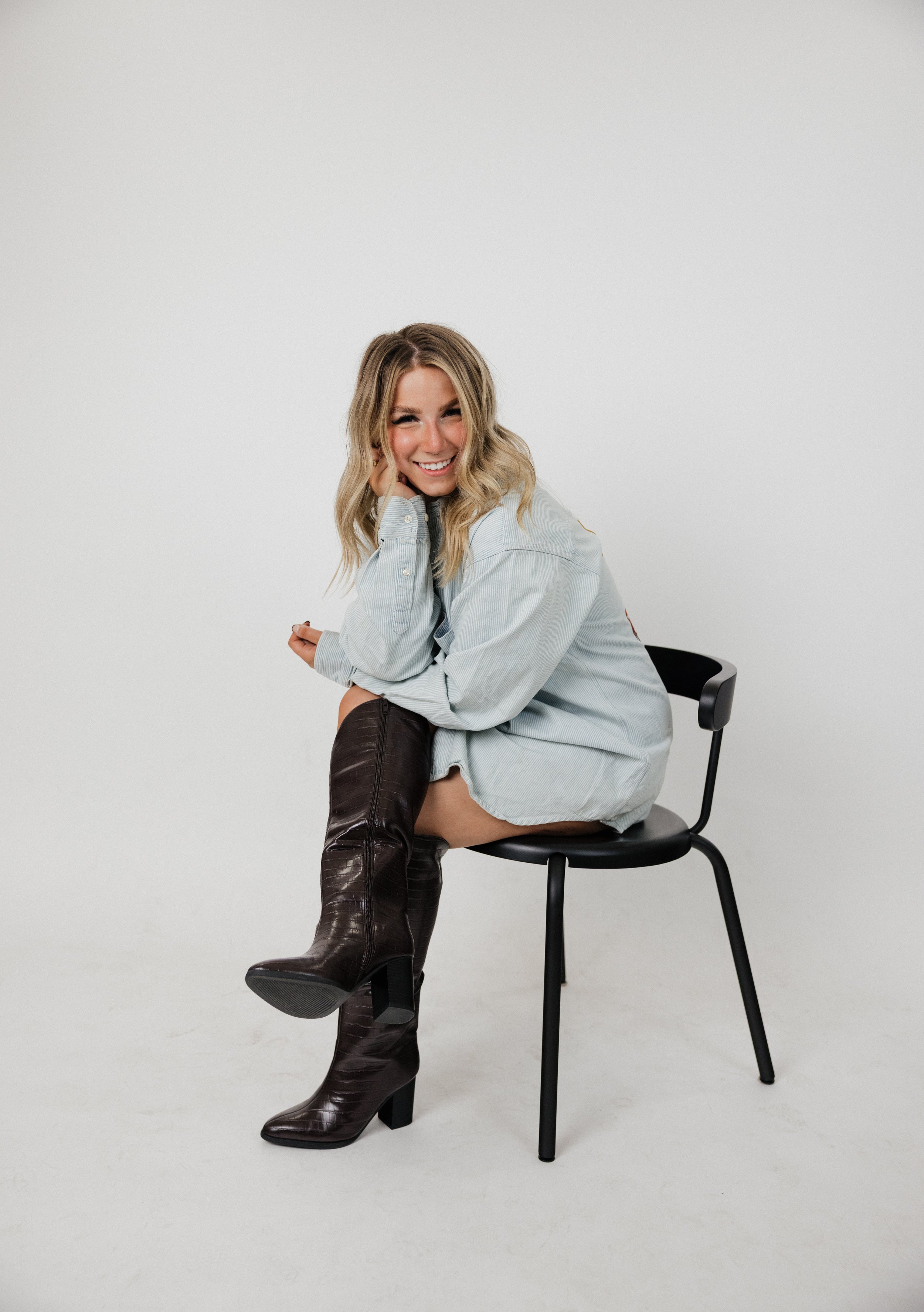 A woman with wavy blonde hair smiling, wearing a light blue button-up shirt and dark brown high-heeled boots, sitting on a chair with one leg crossed over the other against a plain white background.