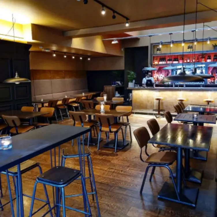 Empty restaurant with wooden floors and tables, some illuminated by candles. Warm lighting, a bar with bottles on shelves, barstools, and modern pendant lights hanging from the ceiling.