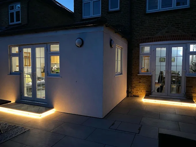 Nighttime view of a house corner with illuminated outdoor lighting along the patio edges, showing two sets of such lights near the doors, with visible windows revealing interior rooms.