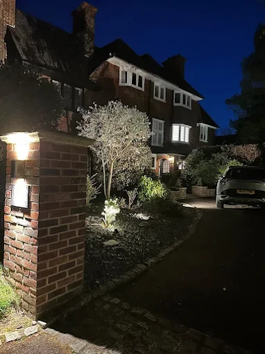 Nighttime view of a residential house with illuminated garden and driveway, brick walls, and a parked car.
