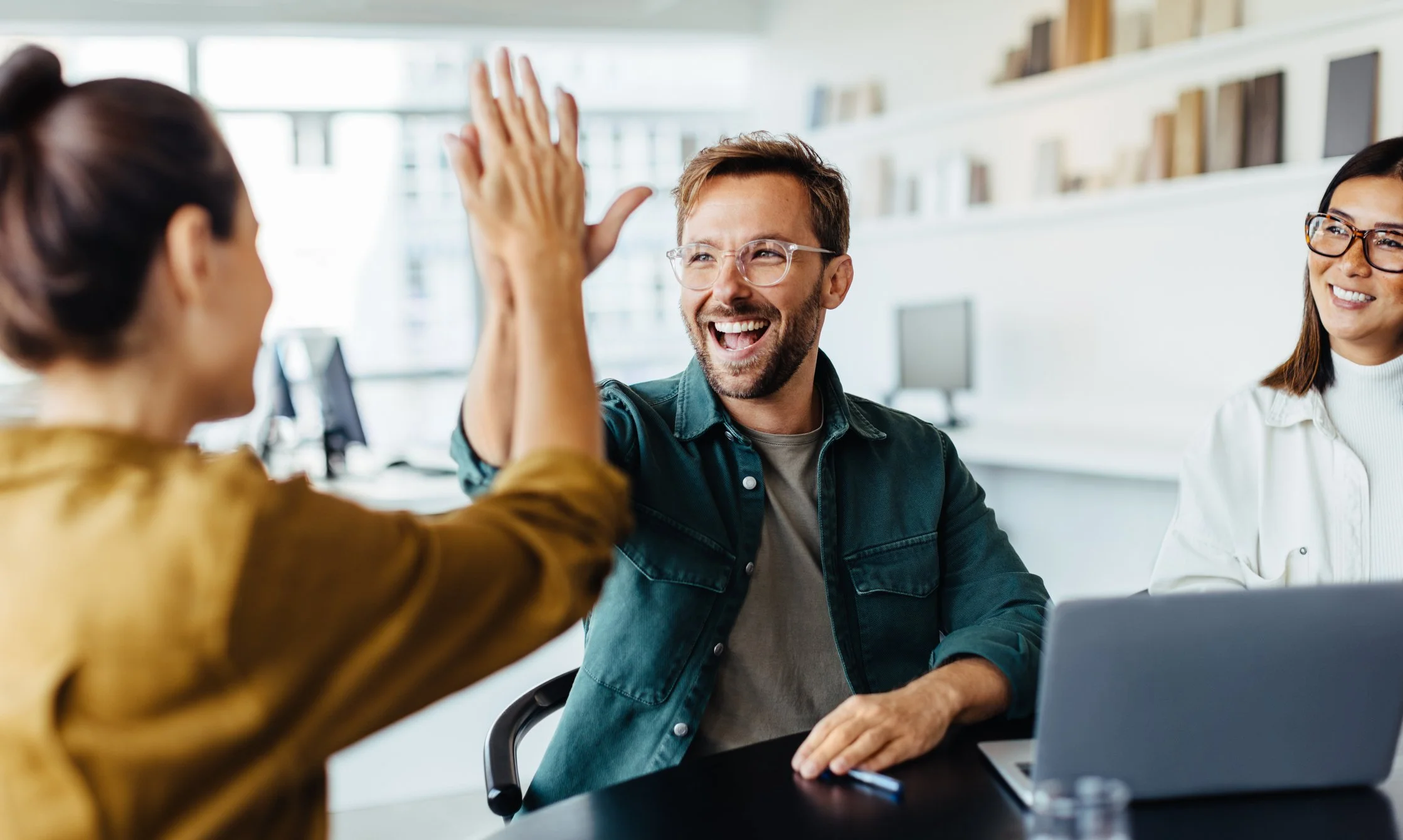 Three smiling people, one man giving a high five to a woman with a laptop on a table in an office setting.