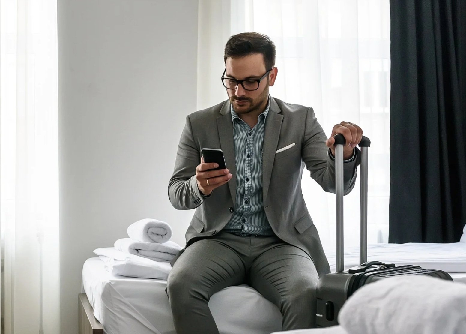 A man in a gray suit sitting on a bed, looking at his phone, with a rolling suitcase beside him. There are folded towels on the bed. The room has light coming through sheer curtains.