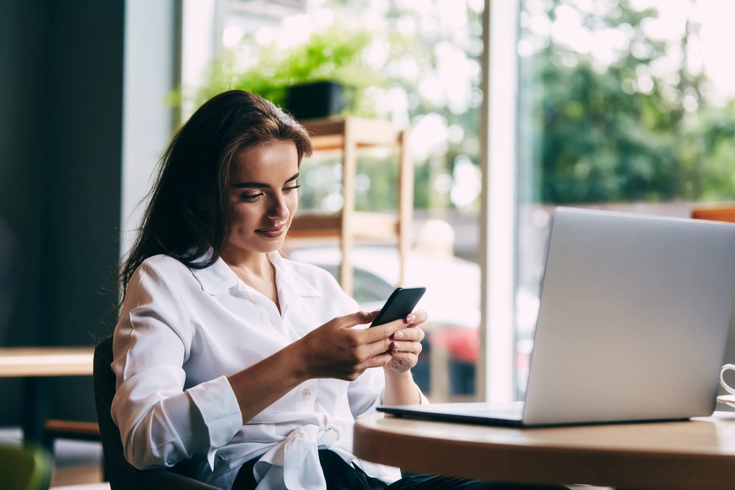 A woman sitting at a wooden table, using her smartphone, with a laptop open in front of her, in a room with large windows and greenery outside.