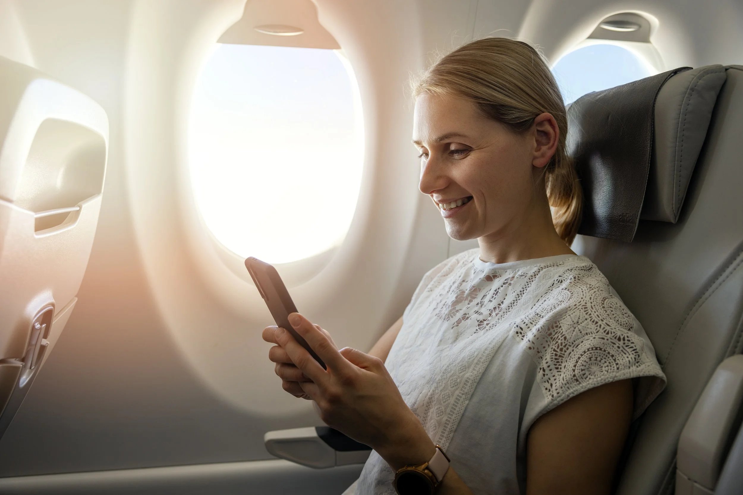 A woman sitting in airplane seat, looking at her phone, smiling, with sunlight coming through the window.