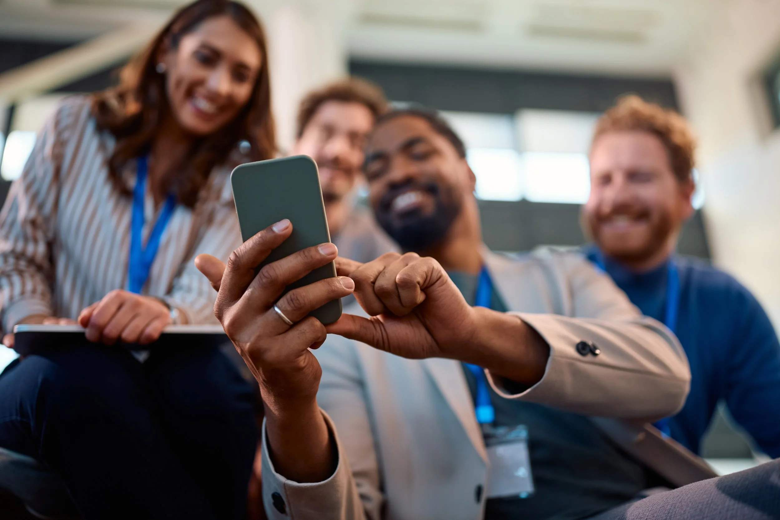 Four people at a conference or event, looking at a smartphone together, smiling, with large windows in the background.