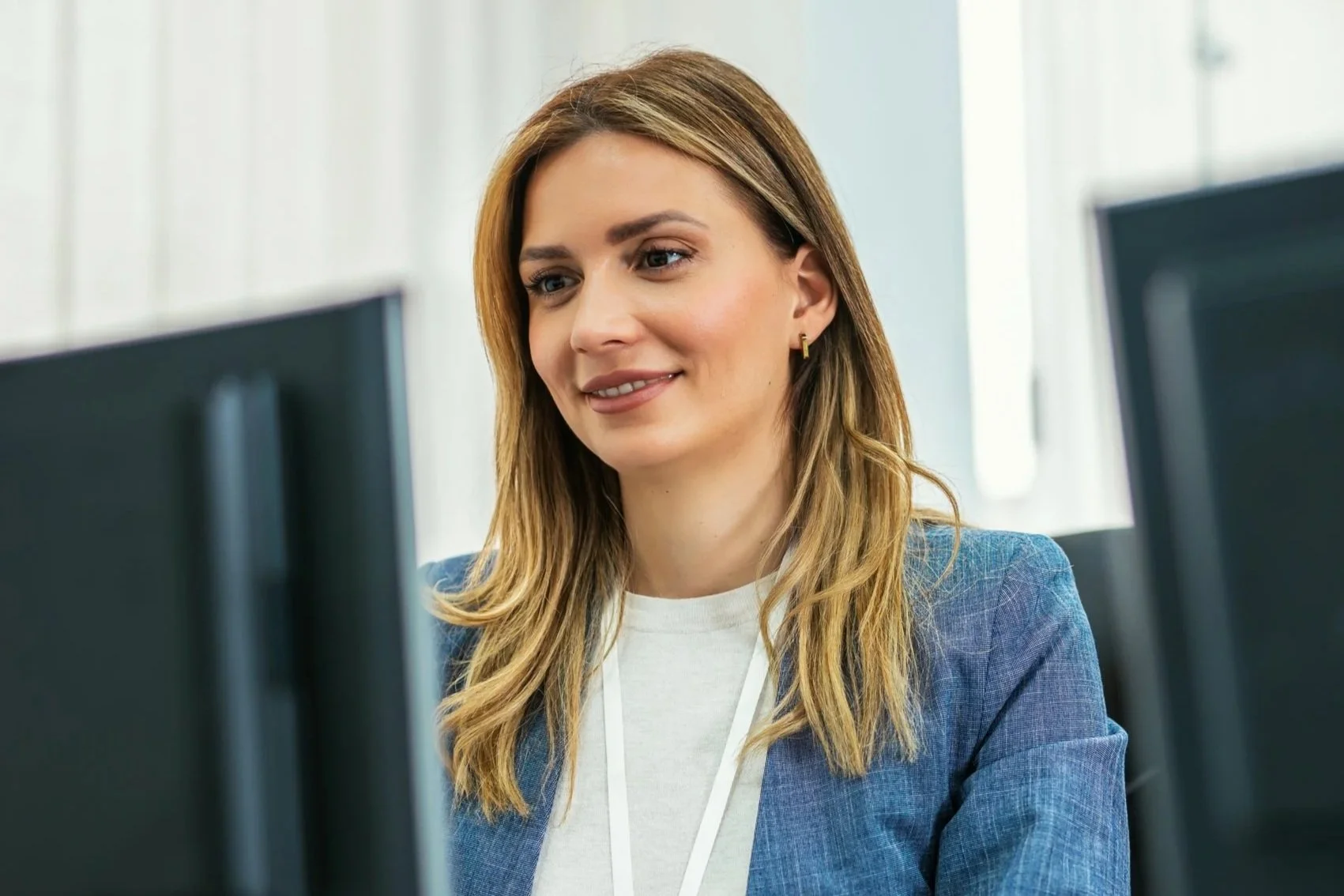 A professional woman with shoulder-length blonde hair, wearing a gray blazer and white top, sitting in front of computer monitors in an office.