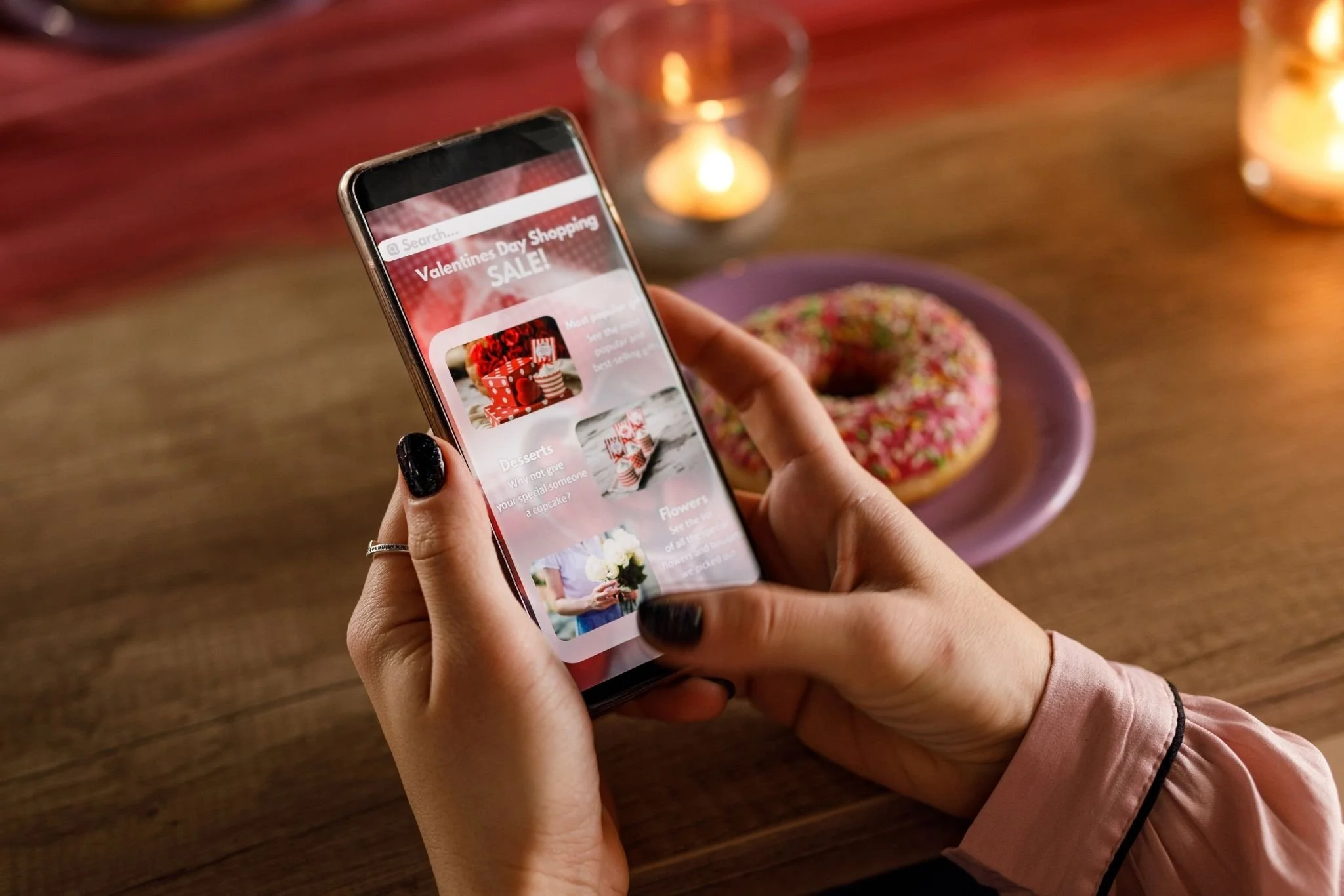 Person holding a smartphone with a Valentine's Day shopping sale app open, with a pink donut on a purple plate and lit candles on a wooden table in the background.