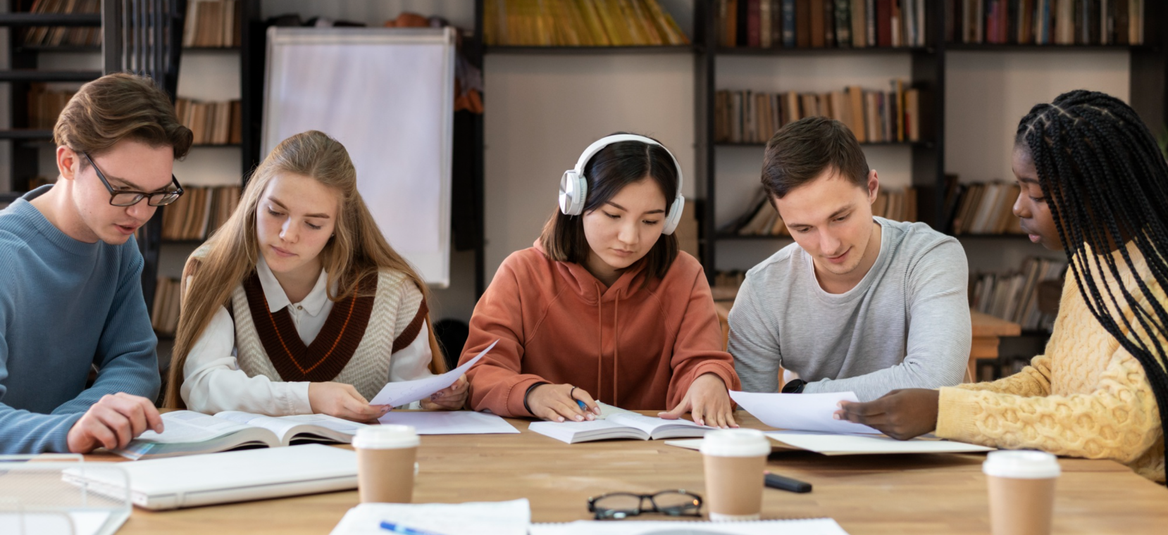 Five young people sitting at a table in a library studying or working on a project with books and papers in front of them.