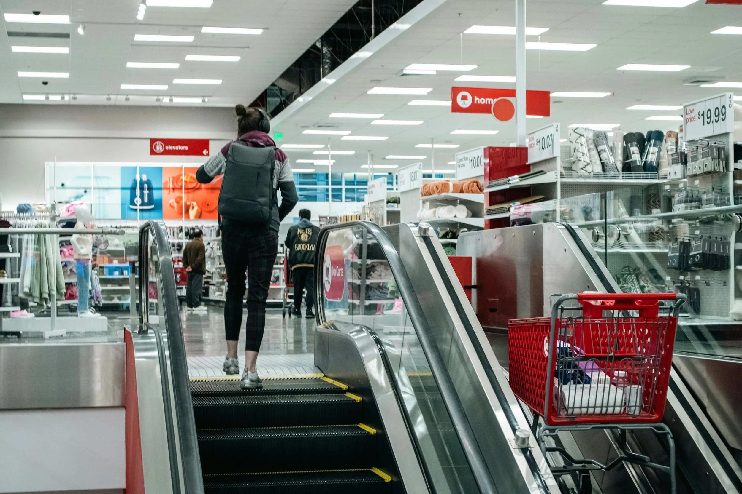 A person with a backpack riding an escalator inside a retail store, with shelves of household items and a shopping cart nearby.