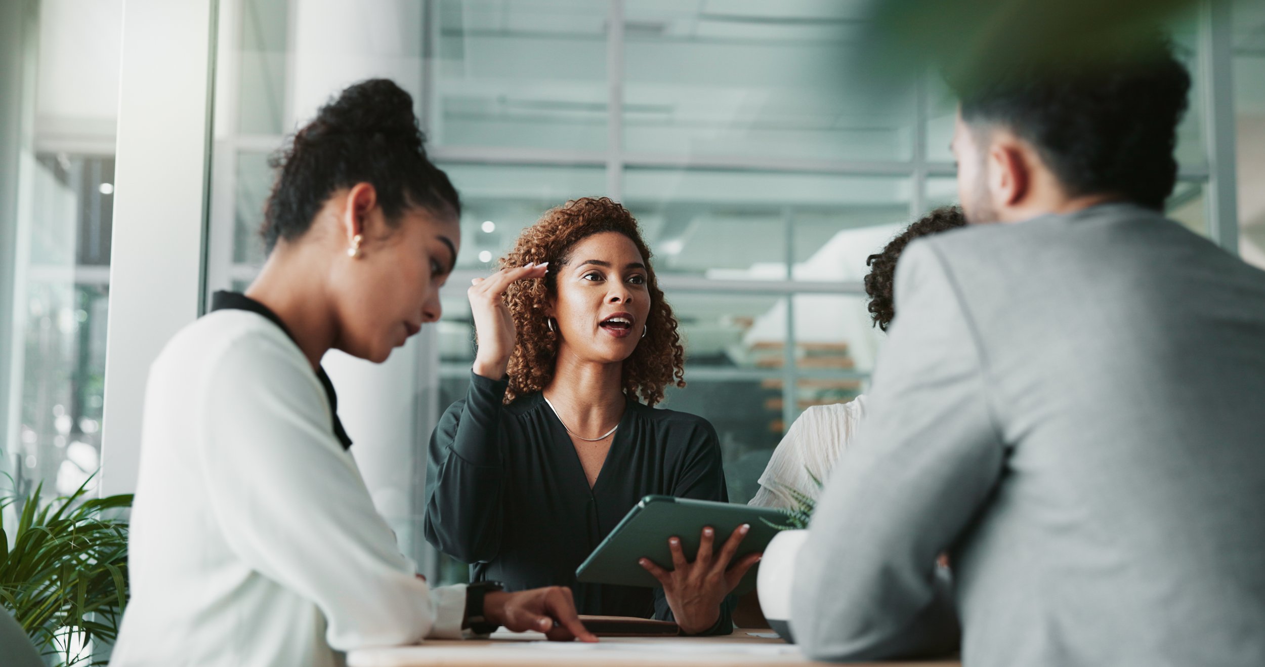 A diverse group of five professionals engaged in a discussion in a modern office space, with one woman speaking and holding a tablet, while others listen attentively.
