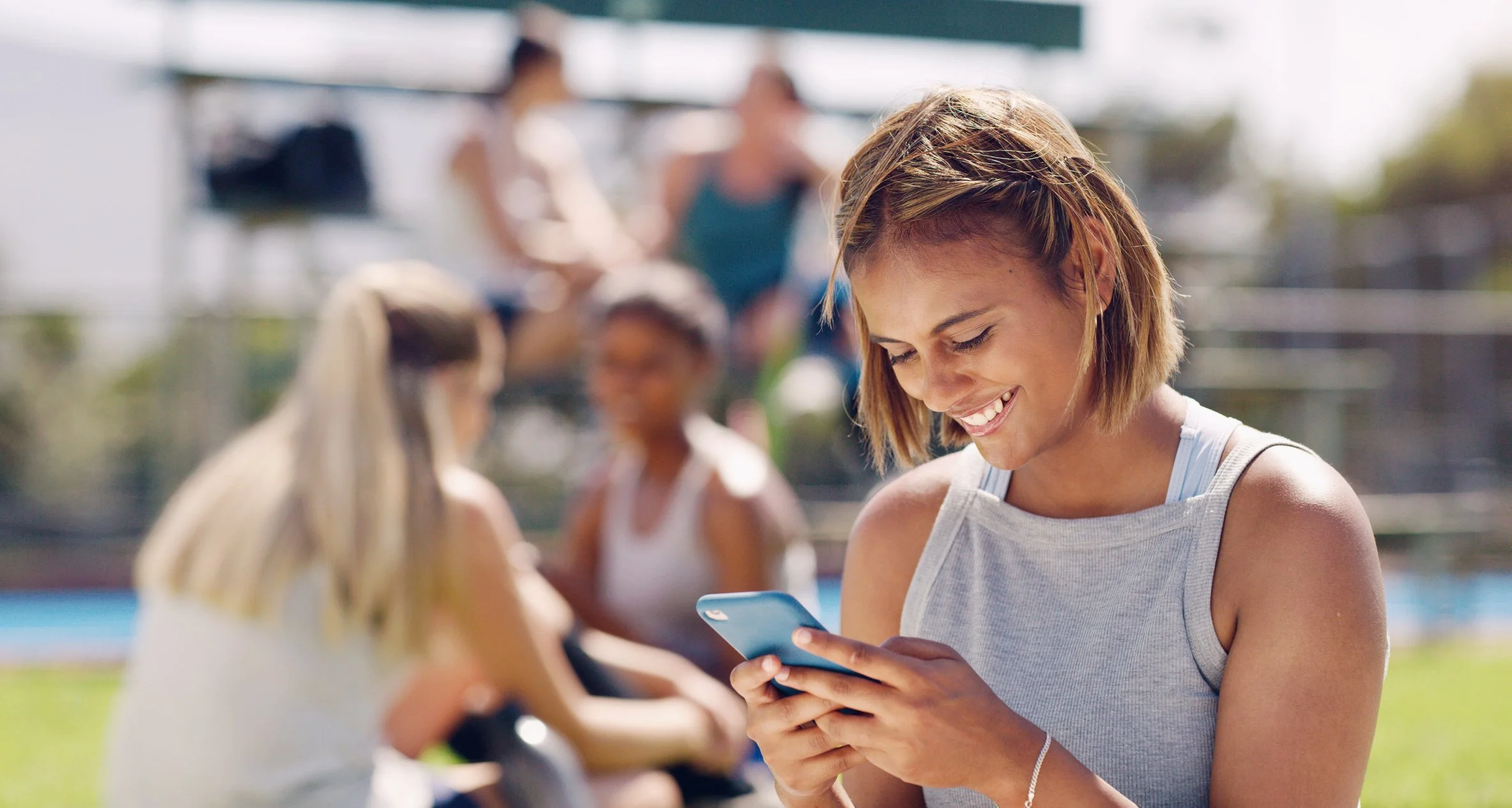 Young woman smiling and looking at her phone outdoors, with other people in the background at a park or recreational area.