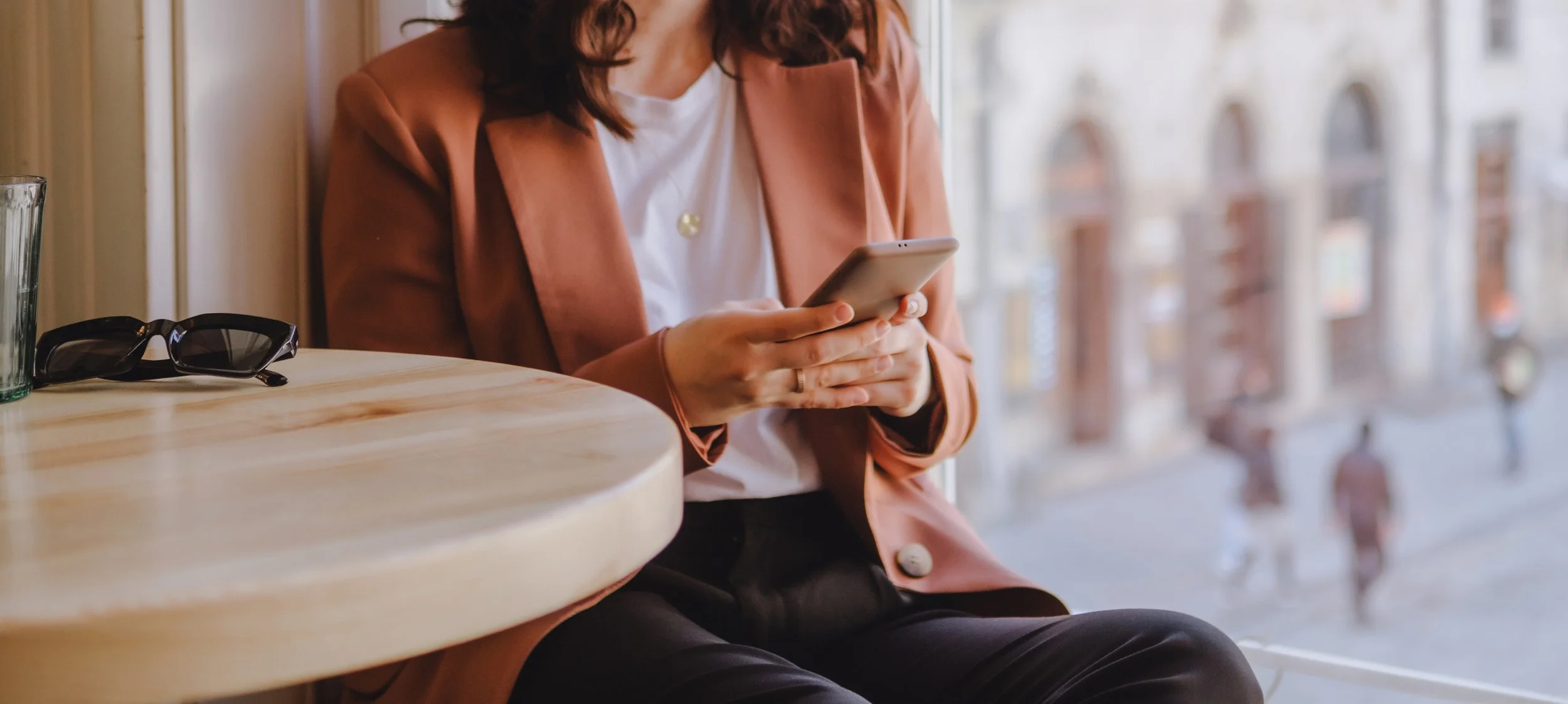 A woman sitting by a window in a café, looking at her smartphone with sunglasses on a table next to her, outside the window is a city street with pedestrians.