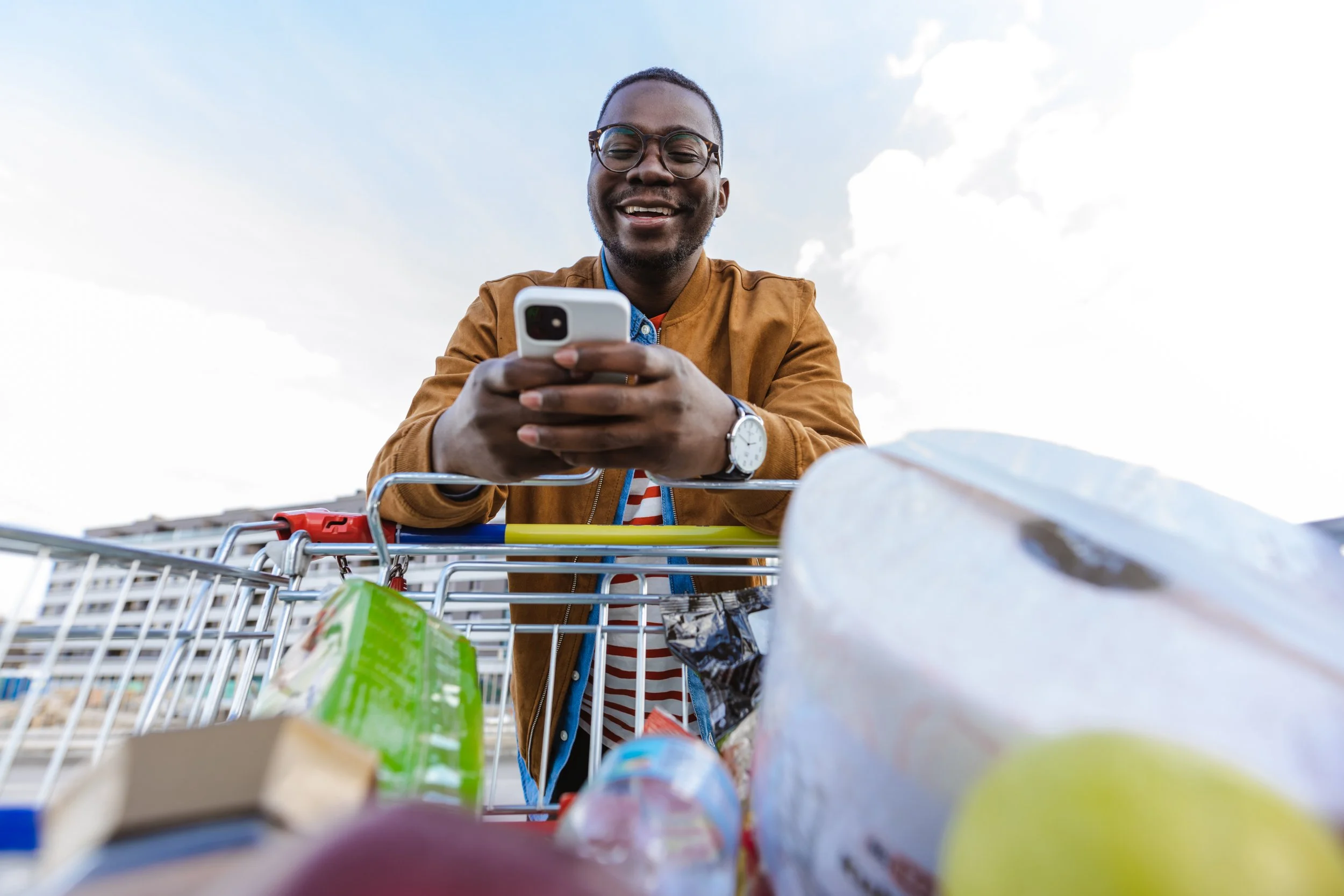 A man smiling and looking at his phone while pushing a shopping cart filled with groceries outdoors.