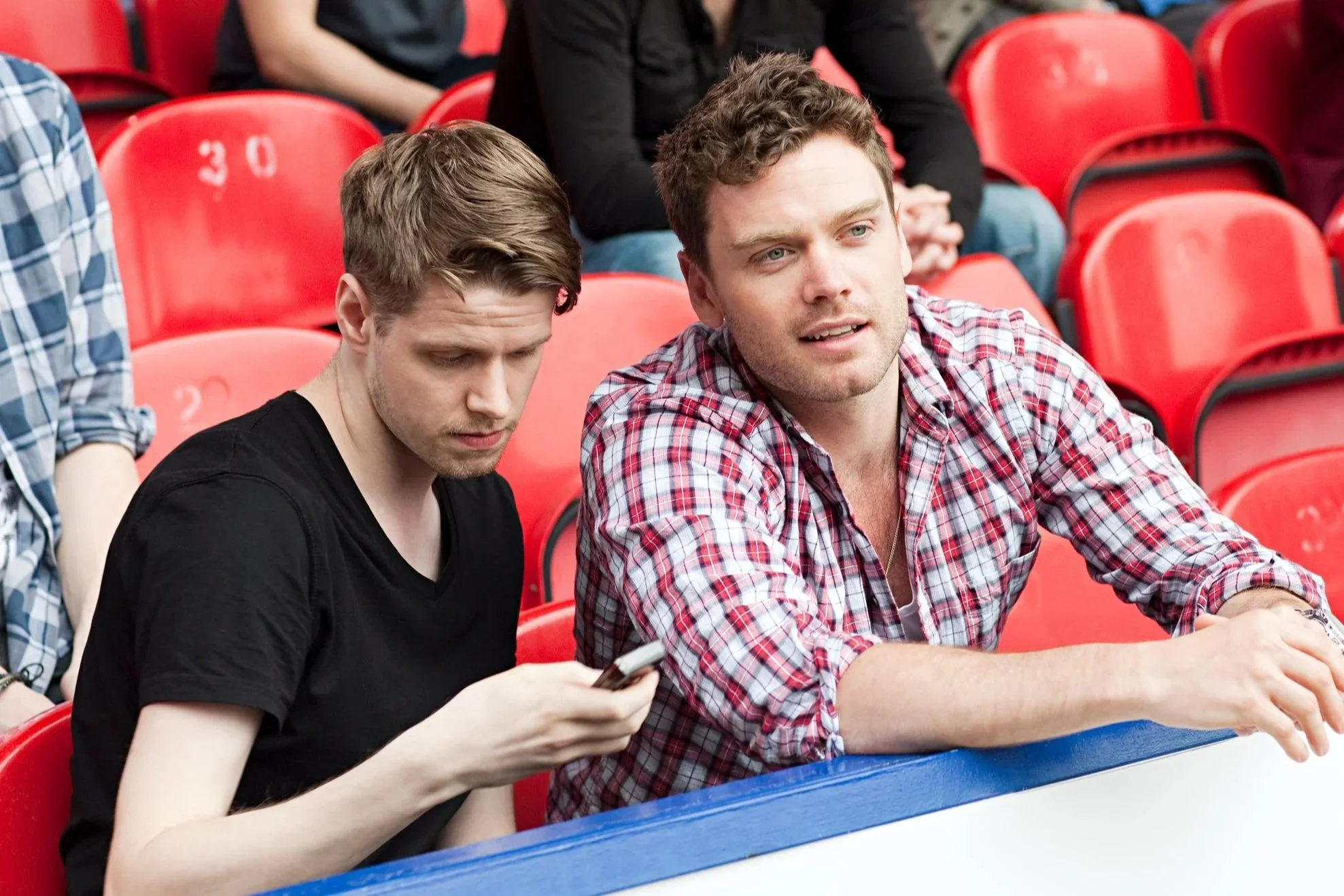 Two young men sitting in red stadium seats; one is looking at his phone, the other is gazing ahead.
