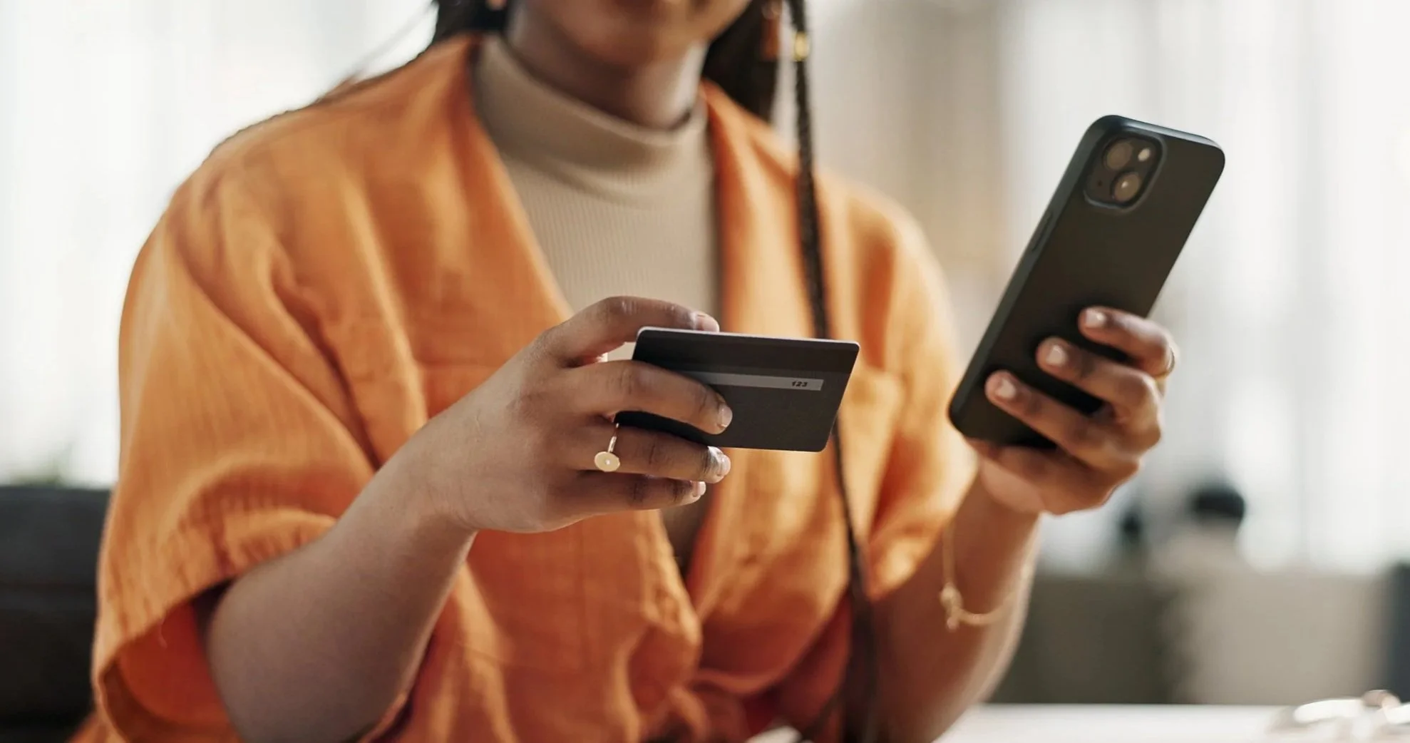 A person in an orange jacket and beige turtleneck holding a credit card in one hand and a smartphone in the other, sitting indoors.