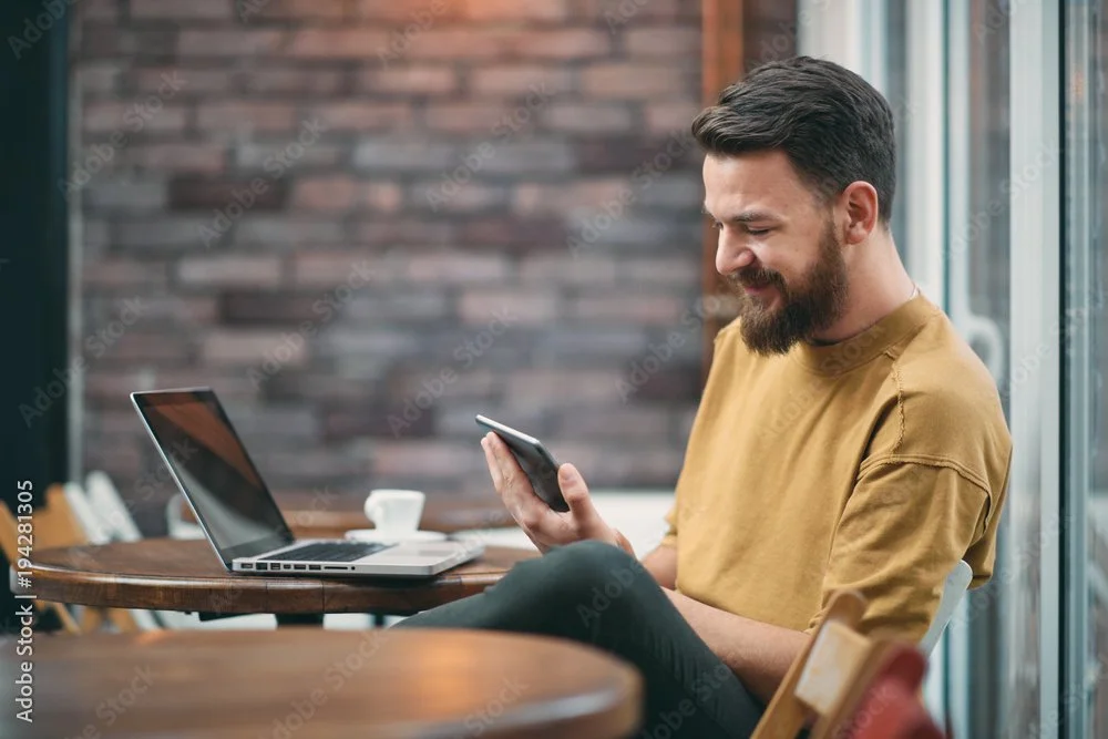 A man with a beard and yellow T-shirt sitting at a table, looking at his phone with a laptop and a cup on the table, in a cozy cafe or home setting.