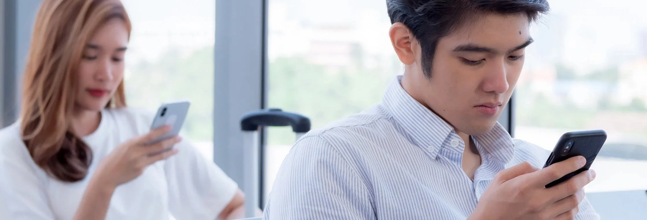 Man and woman sitting near large windows, both using smartphones, with a blurred background of cityscape.