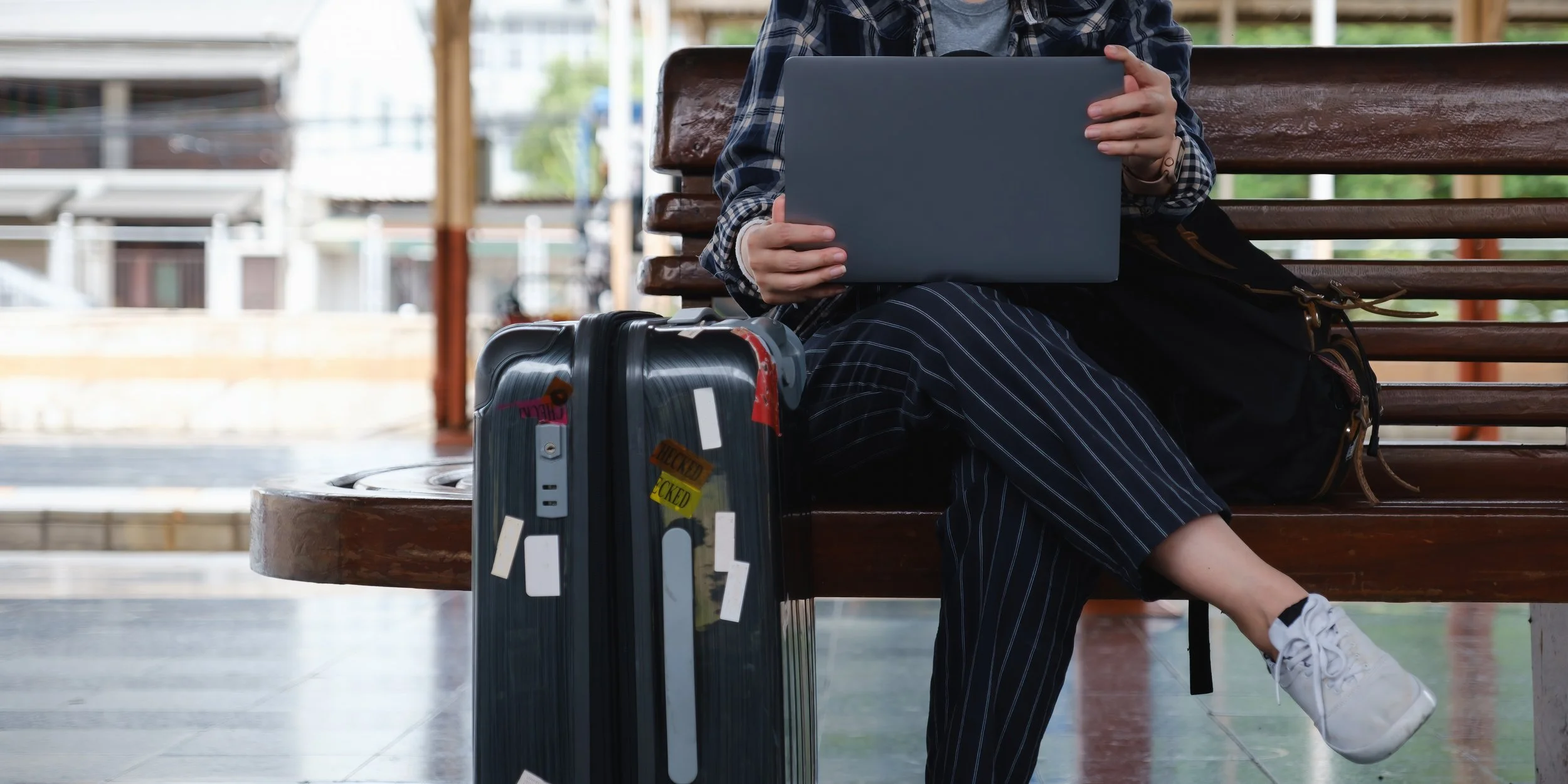 A person sitting on a bench in a train station, using a laptop with a backpack nearby and a suitcase with stickers, wearing sneakers and striped pants.