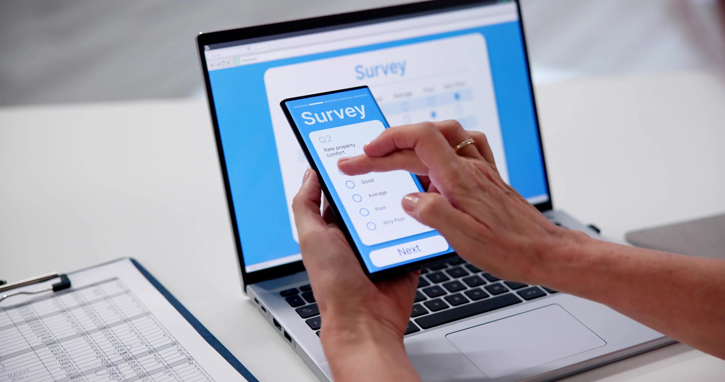 Person holding a smartphone with a survey on the screen, in front of a laptop displaying a survey webpage, and a spreadsheet on the side.