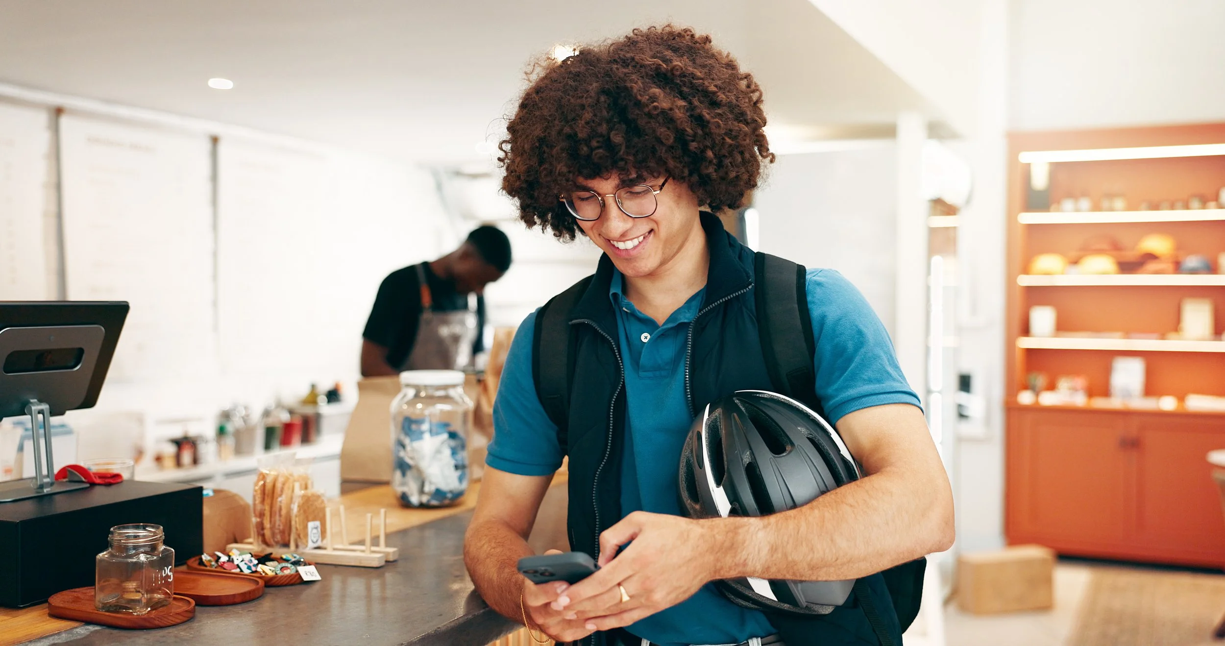 A young man with curly hair and glasses at a cafe counter, holding a bike helmet and using his phone, smiling.