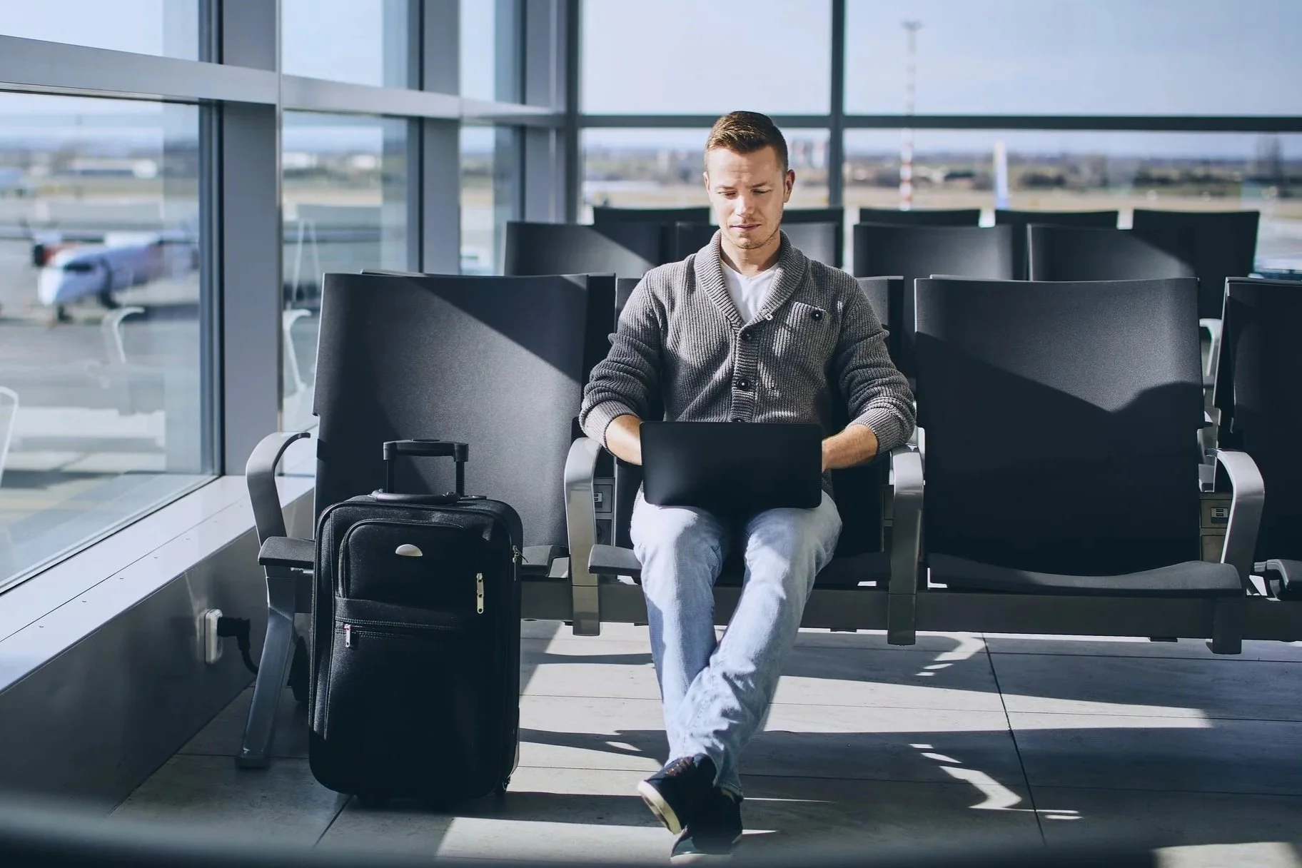 A young man sitting in an airport terminal, using a laptop, with a suitcase beside him, looking at the screen, near large windows with an airport tarmac outside.