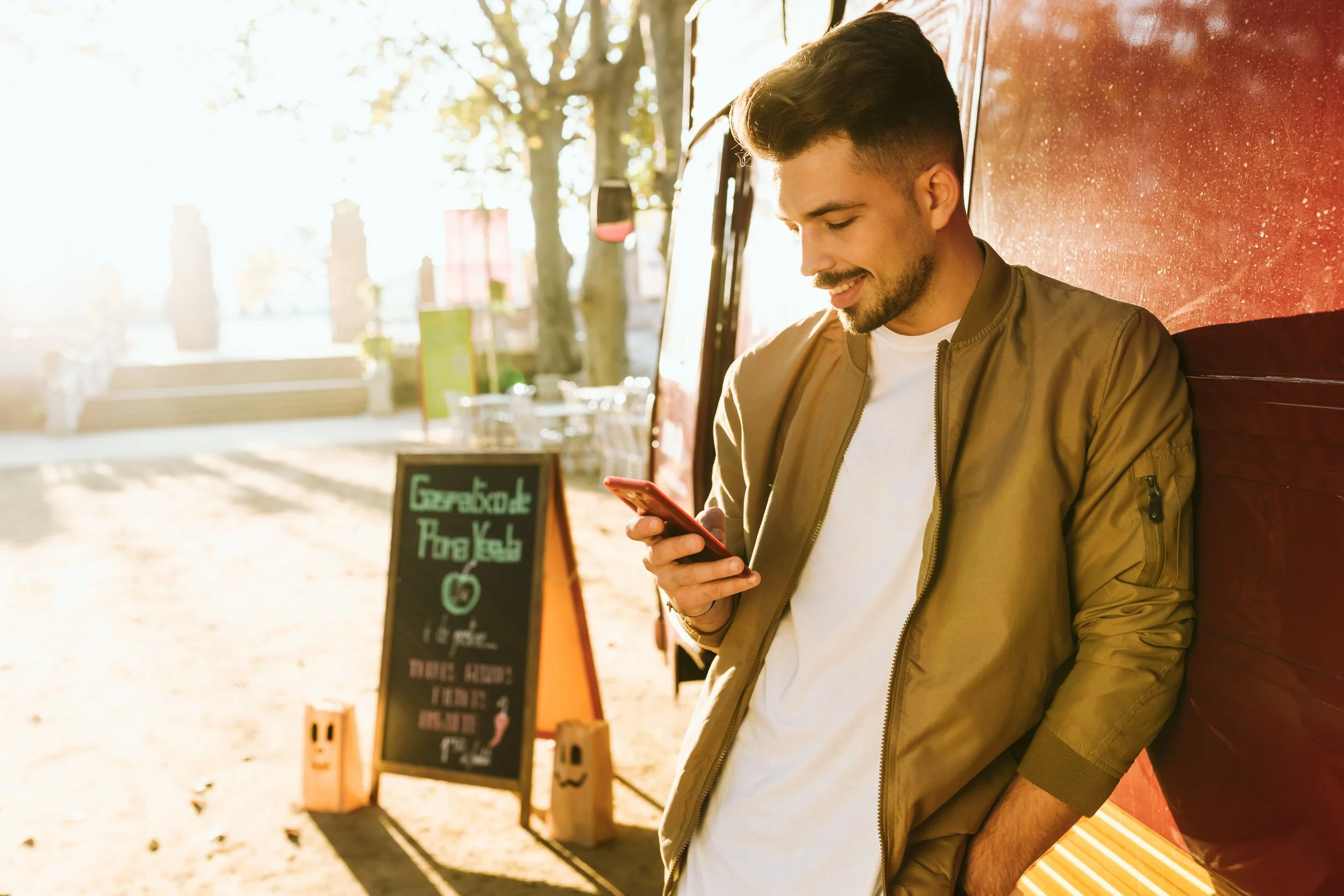 A young man standing against a red food truck, smiling while looking at his phone, with a chalkboard sign for juice in the background on a sunny autumn day.