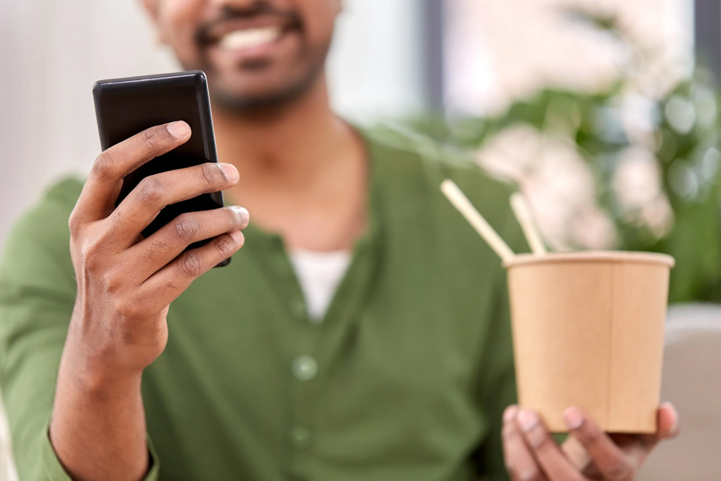 A man holding a takeout container with two chopsticks and using his smartphone with a blurred background.