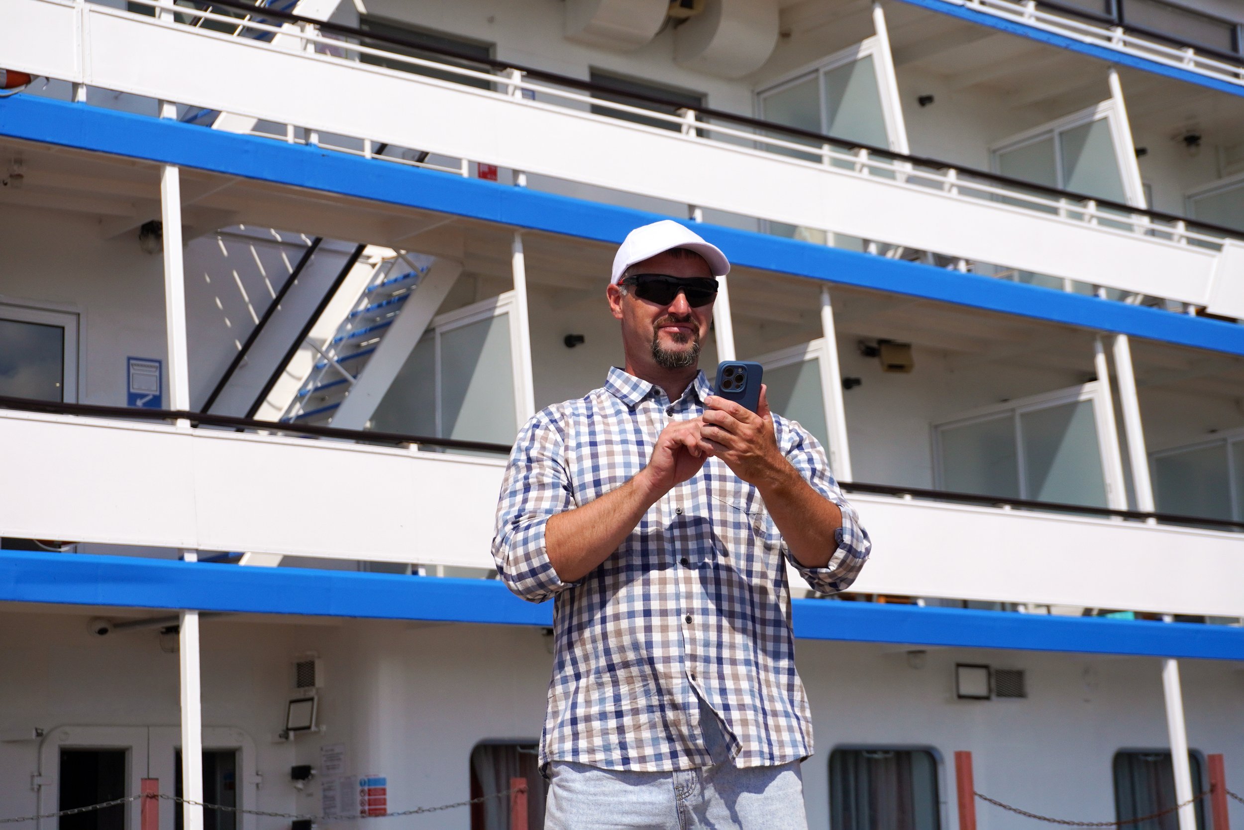 A man wearing sunglasses, a white cap, and a checkered shirt is using his cellphone outdoors, in front of a boat or cruise ship with multiple decks and balconies.