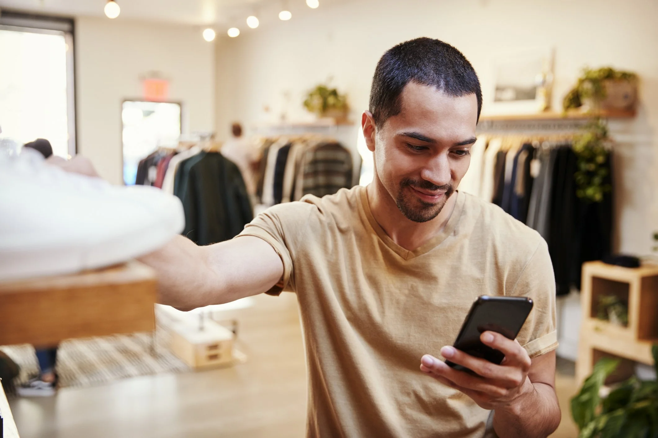 Man shopping in store looking at smartphone with clothing racks in background.