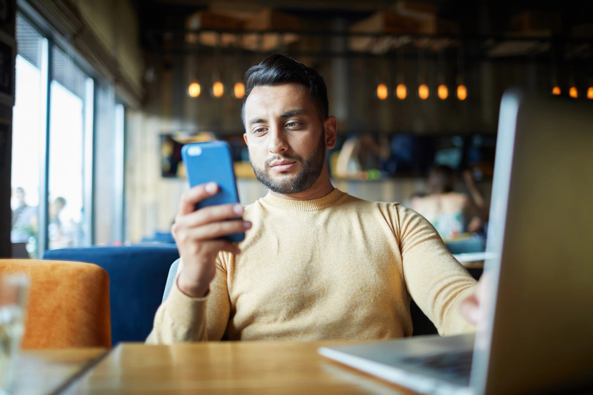 A man sitting in a cafe looking at his smartphone with a laptop open in front of him.