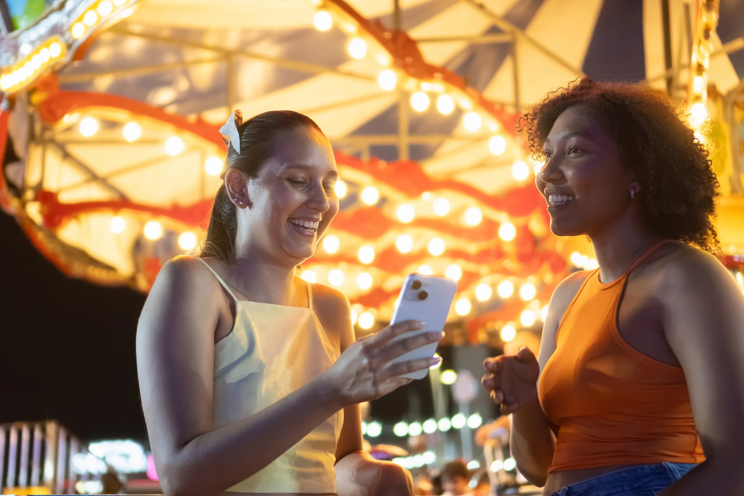 Two women smiling and looking at a smartphone at a night carnival with bright lights and a carousel in the background.