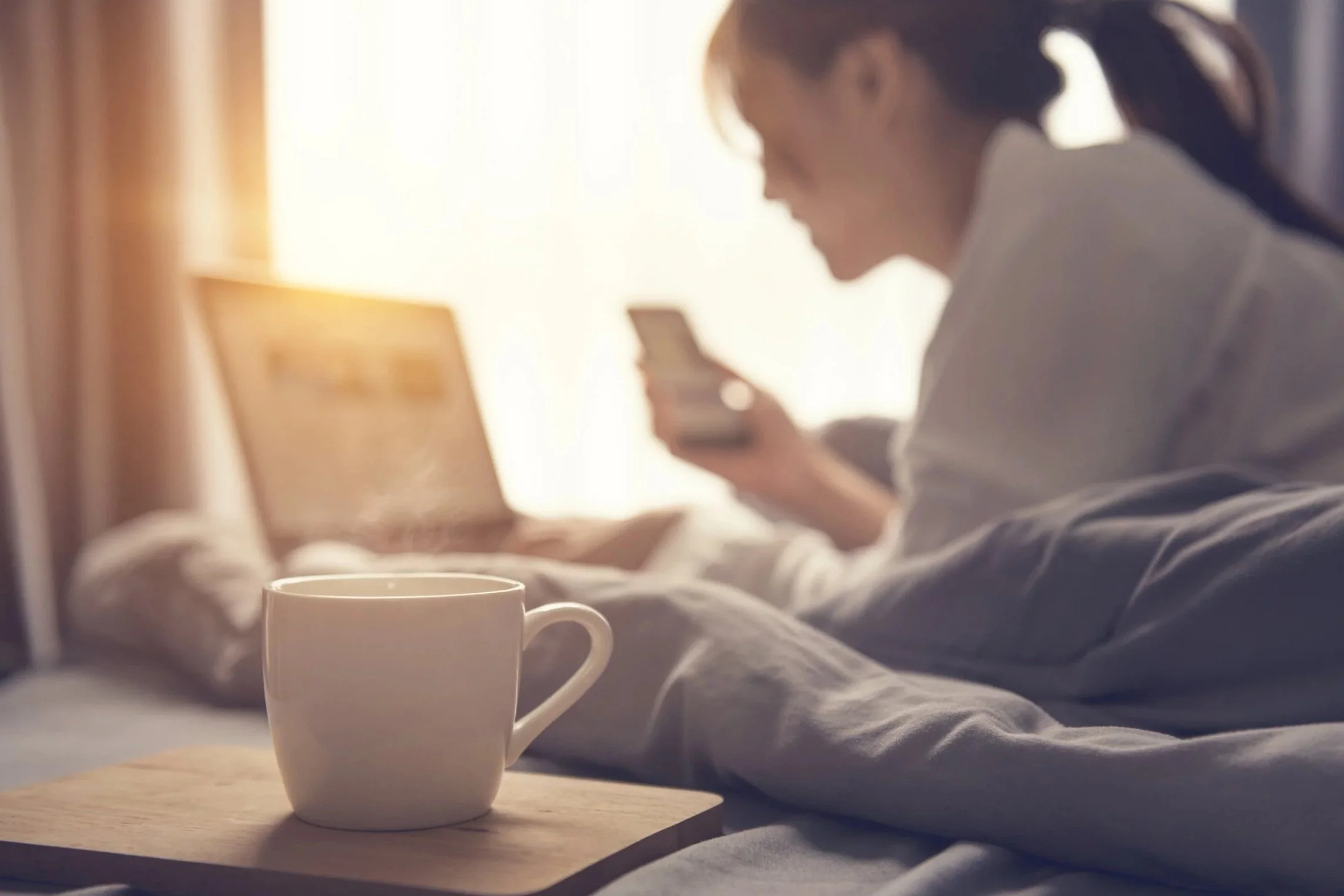 A woman sitting on a bed using her phone with a laptop in front of her and a white mug on a wooden tray in the foreground, in a sunlit room.