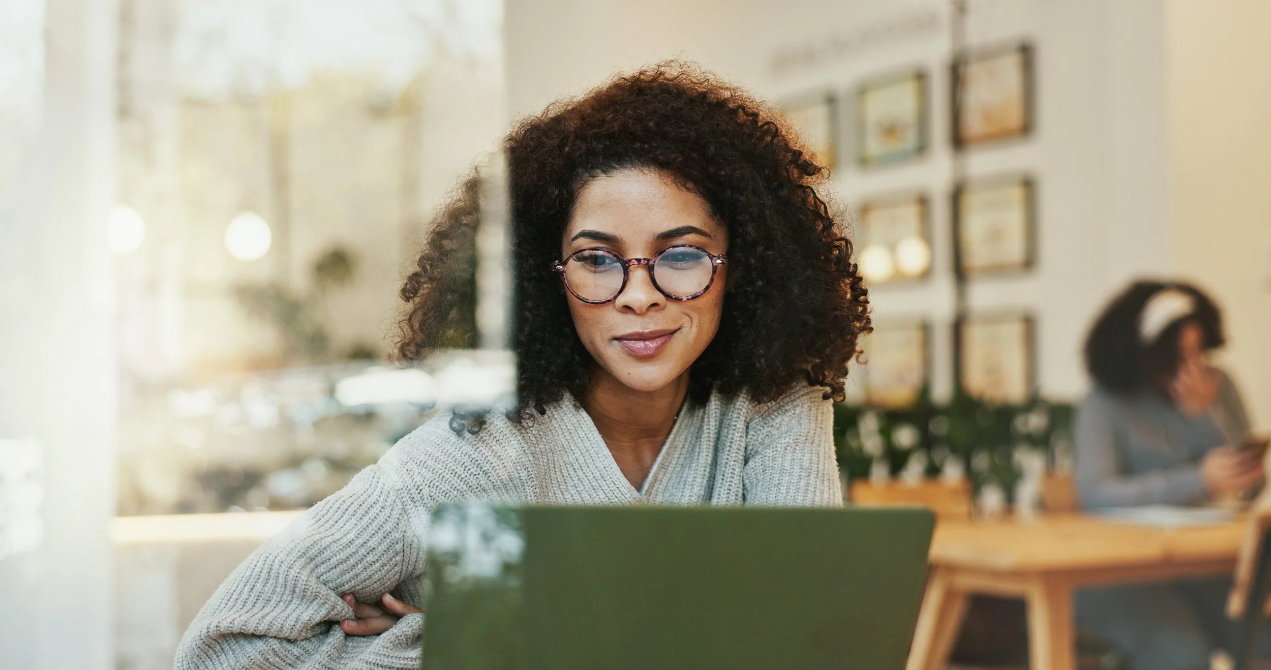 A woman with curly hair and glasses working on a laptop in a cafe, with another person in the background using a phone.