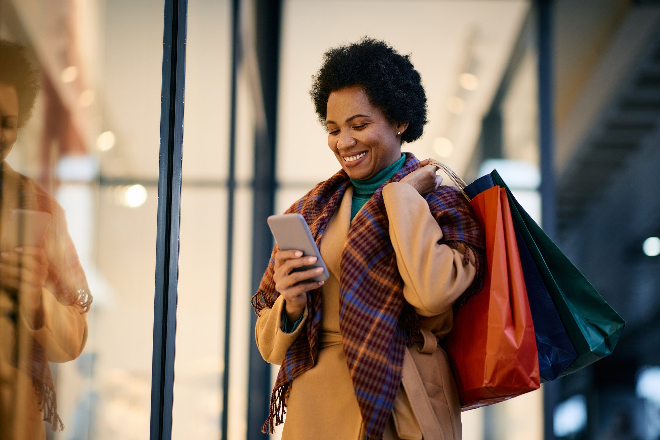 Woman in beige coat holding shopping bags, using smartphone, inside a modern shopping mall.