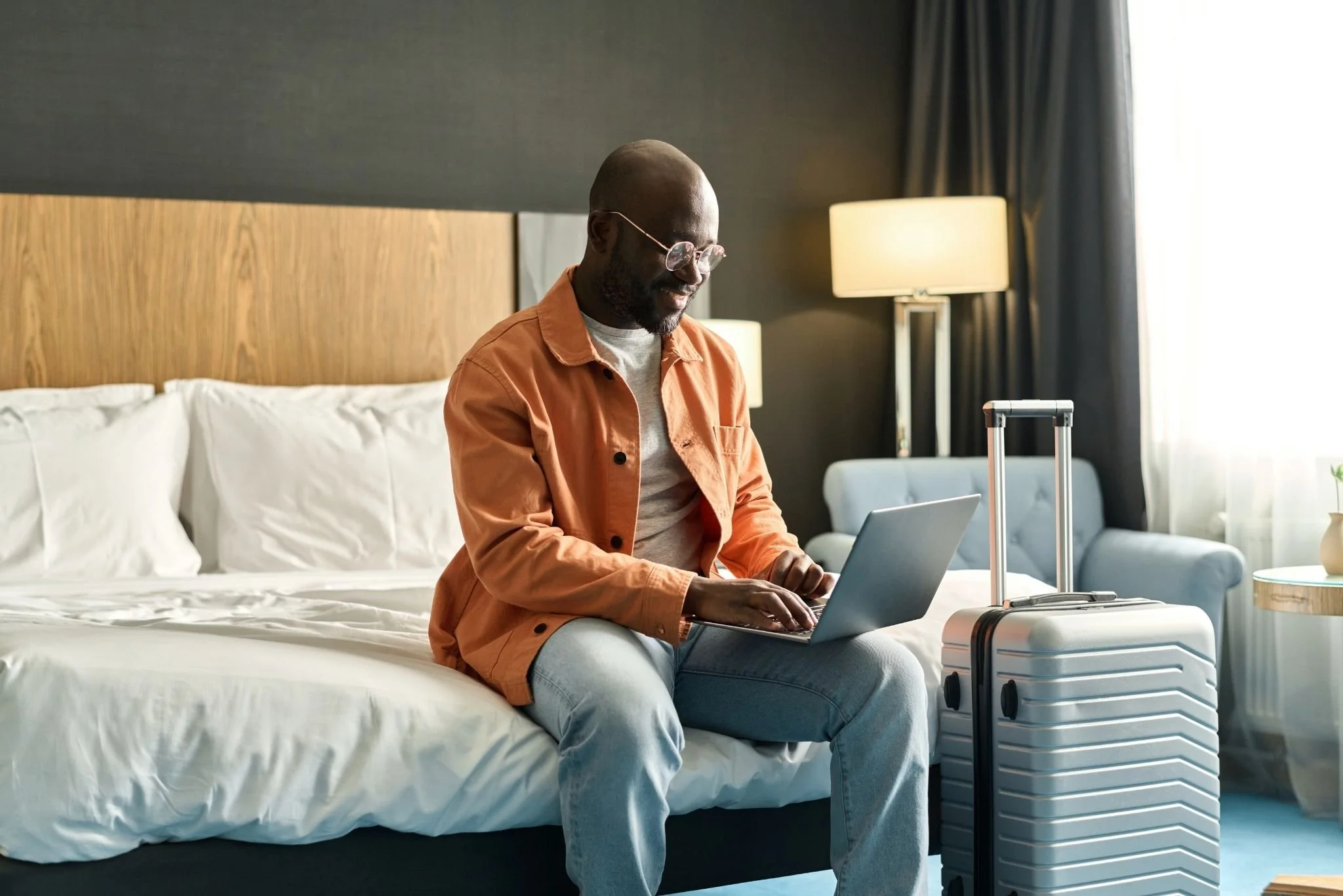 Man sitting on hotel bed using laptop with suitcase beside him in hotel room.