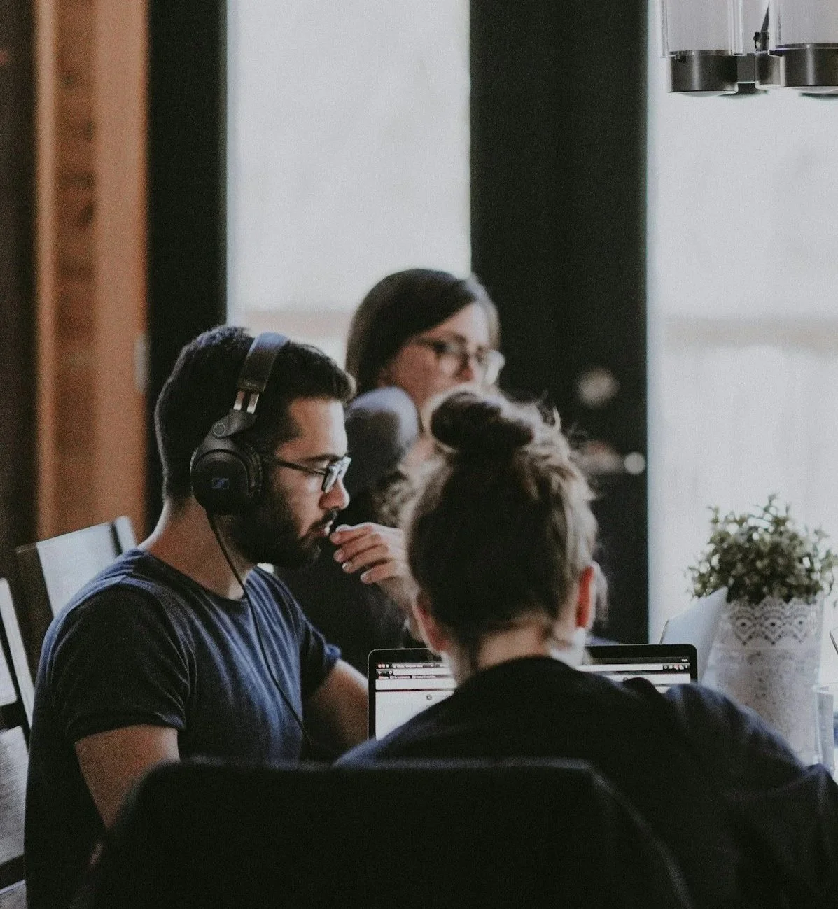 Three people sitting at a table, working on laptops. One person wears glasses and headphones, another has their hair tied up, and a third person with glasses is slightly blurred in the background. There is a vase with flowers on the table.