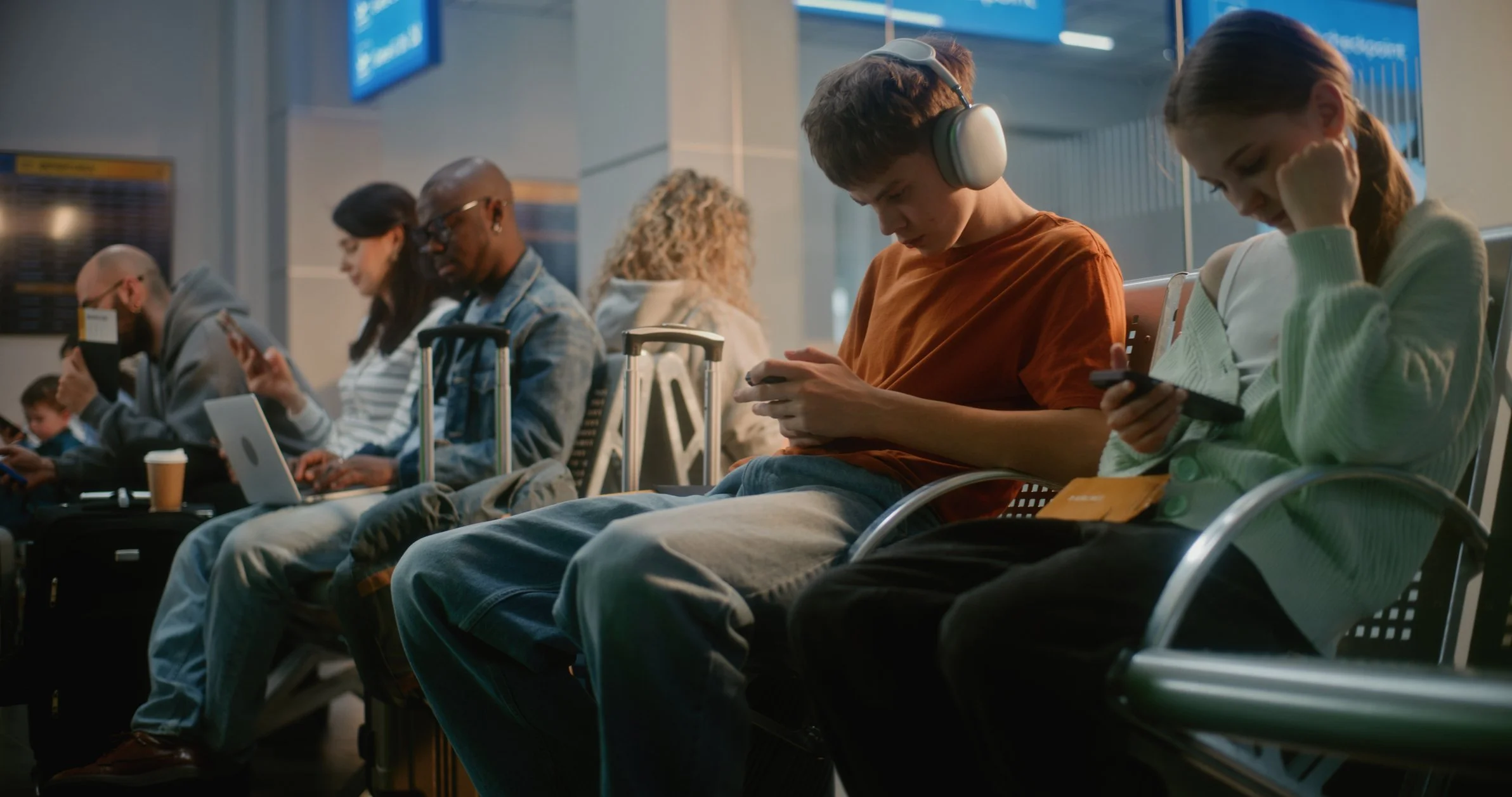 People sitting in an airport waiting area, engrossed in their devices, with luggage and laptops nearby.