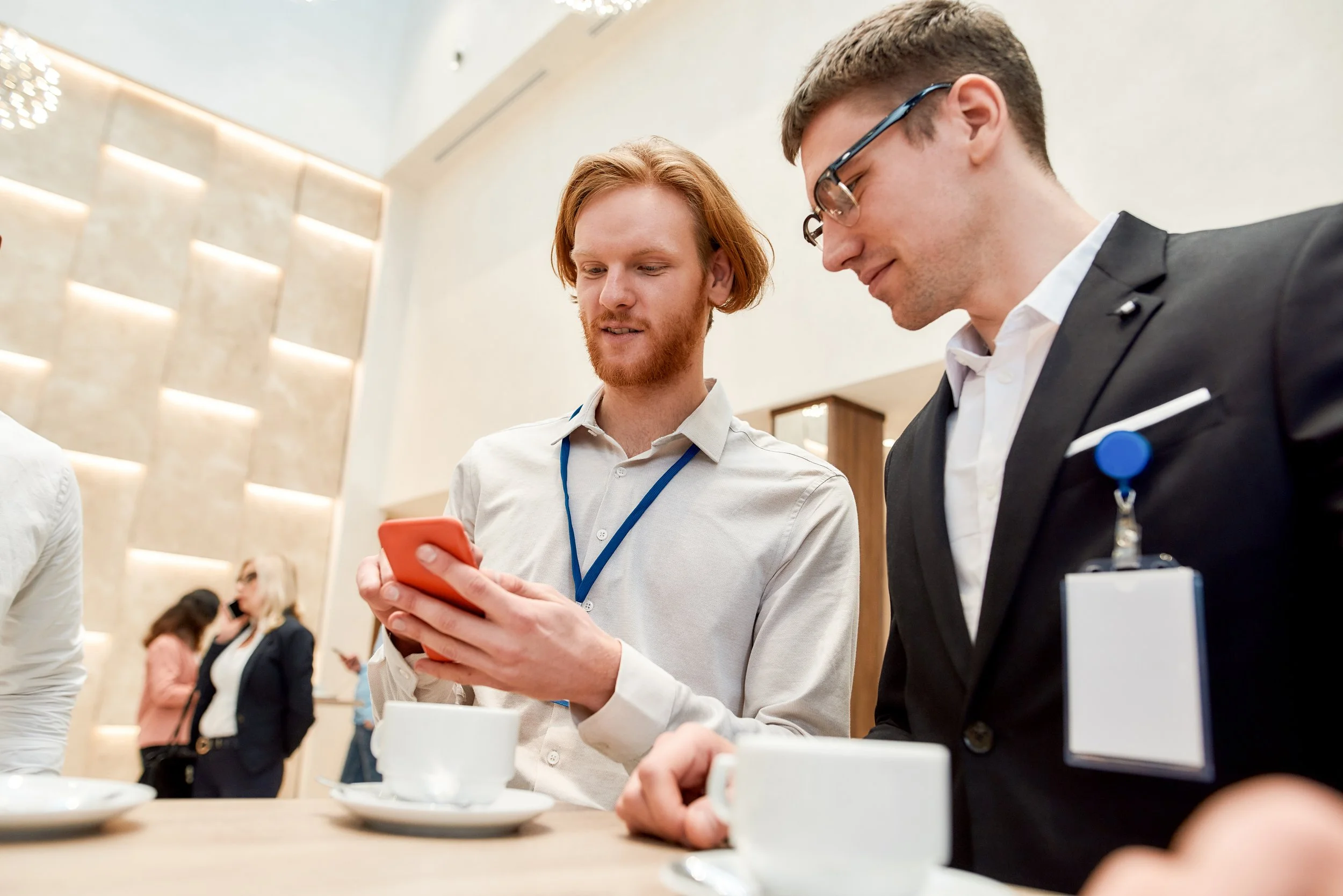 Two men in business attire are standing at a table with coffee cups, looking at a smartphone together, in a modern conference setting.