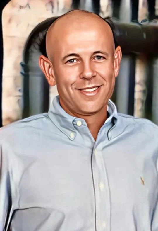 A man with a bald head and light skin, smiling, wearing a light blue button-up shirt, with an outdoor background featuring a brick wall and black railings.