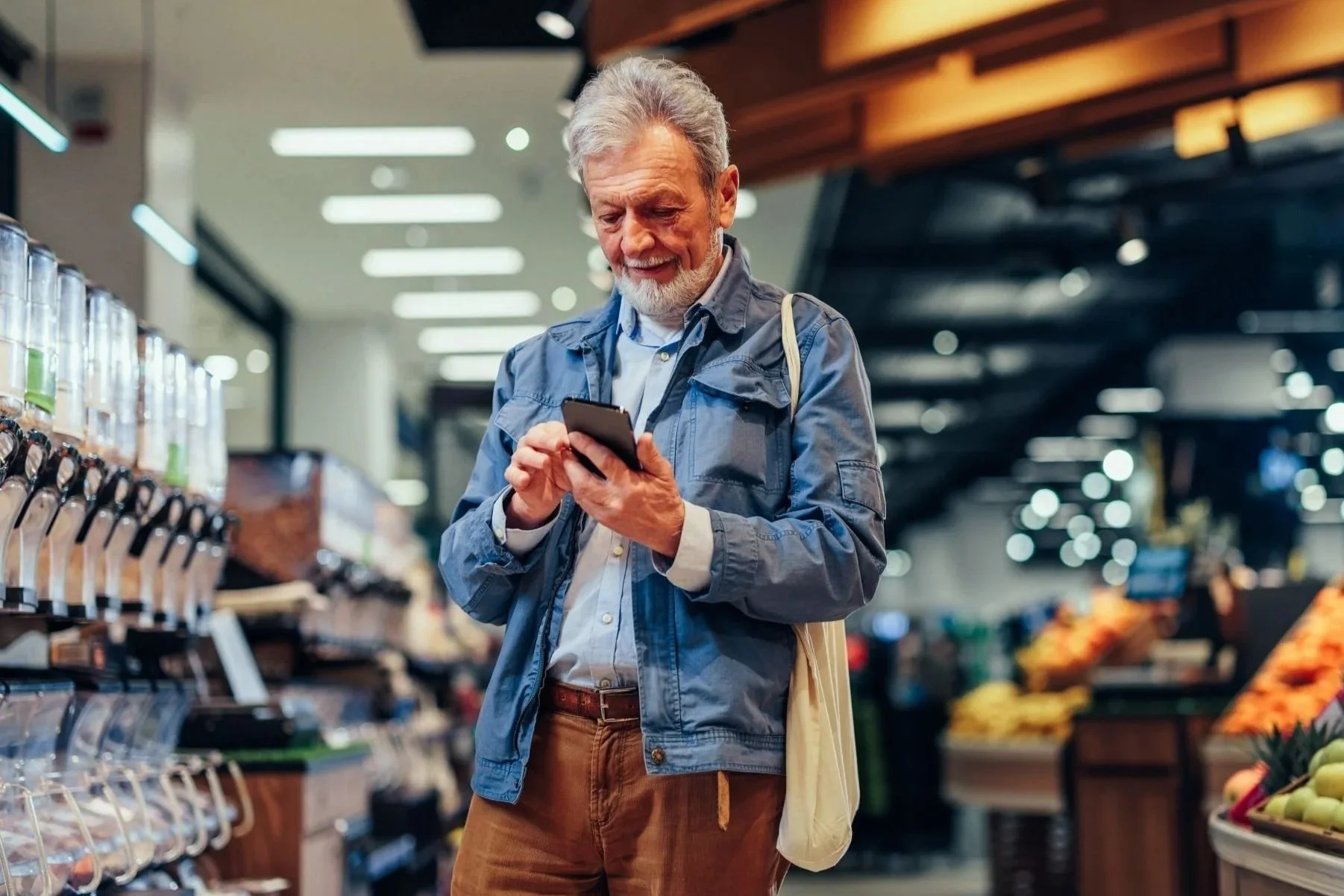 An elderly man with gray hair and beard, wearing a blue jacket and brown pants, shopping in a grocery store while using his smartphone.