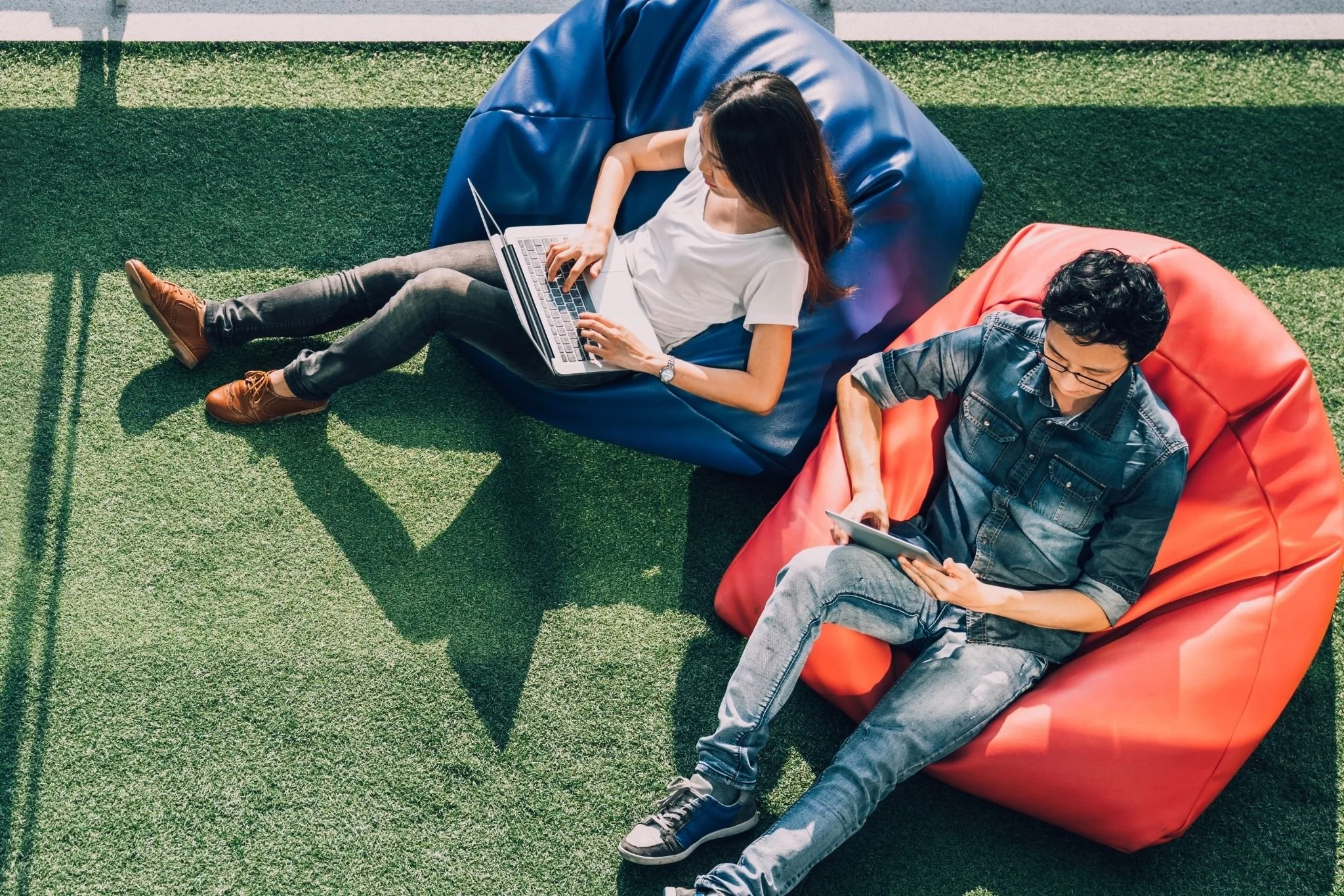 Two young people are relaxing on bean bags on an outdoor grassy area, working on their electronic devices. The woman on the left is using a laptop while the man on the right is using a tablet.