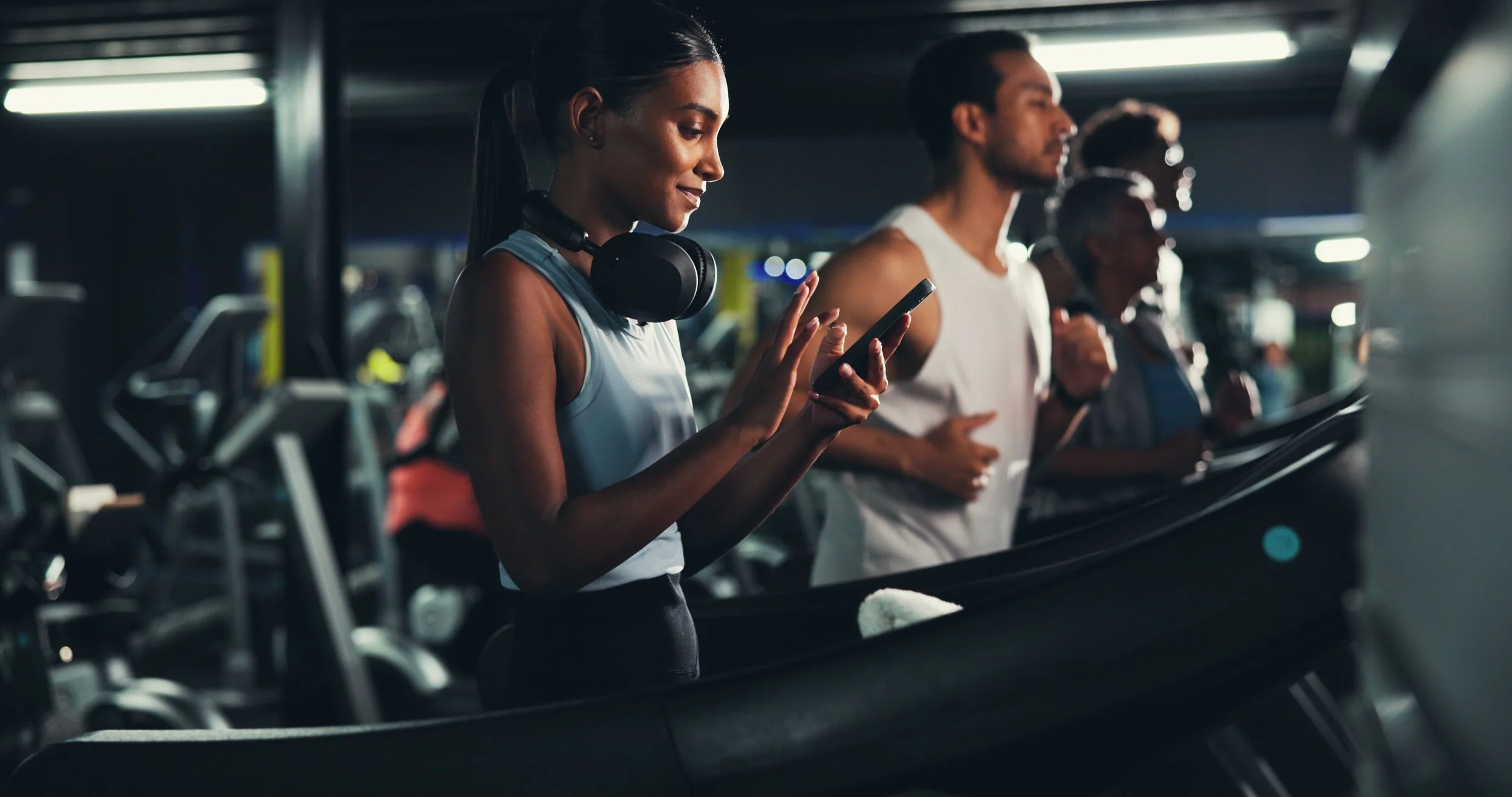 People at a gym using treadmills, with a woman in the foreground looking at her phone and wearing headphones.