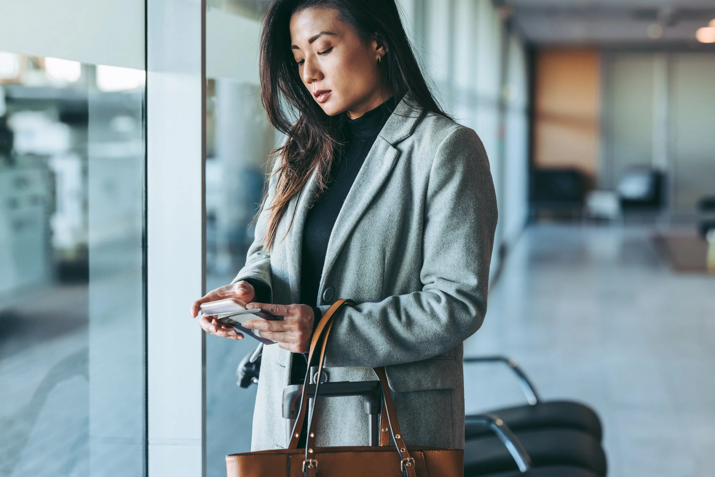 A woman in a gray blazer standing at an airport terminal, looking at her phone while holding a brown handbag.