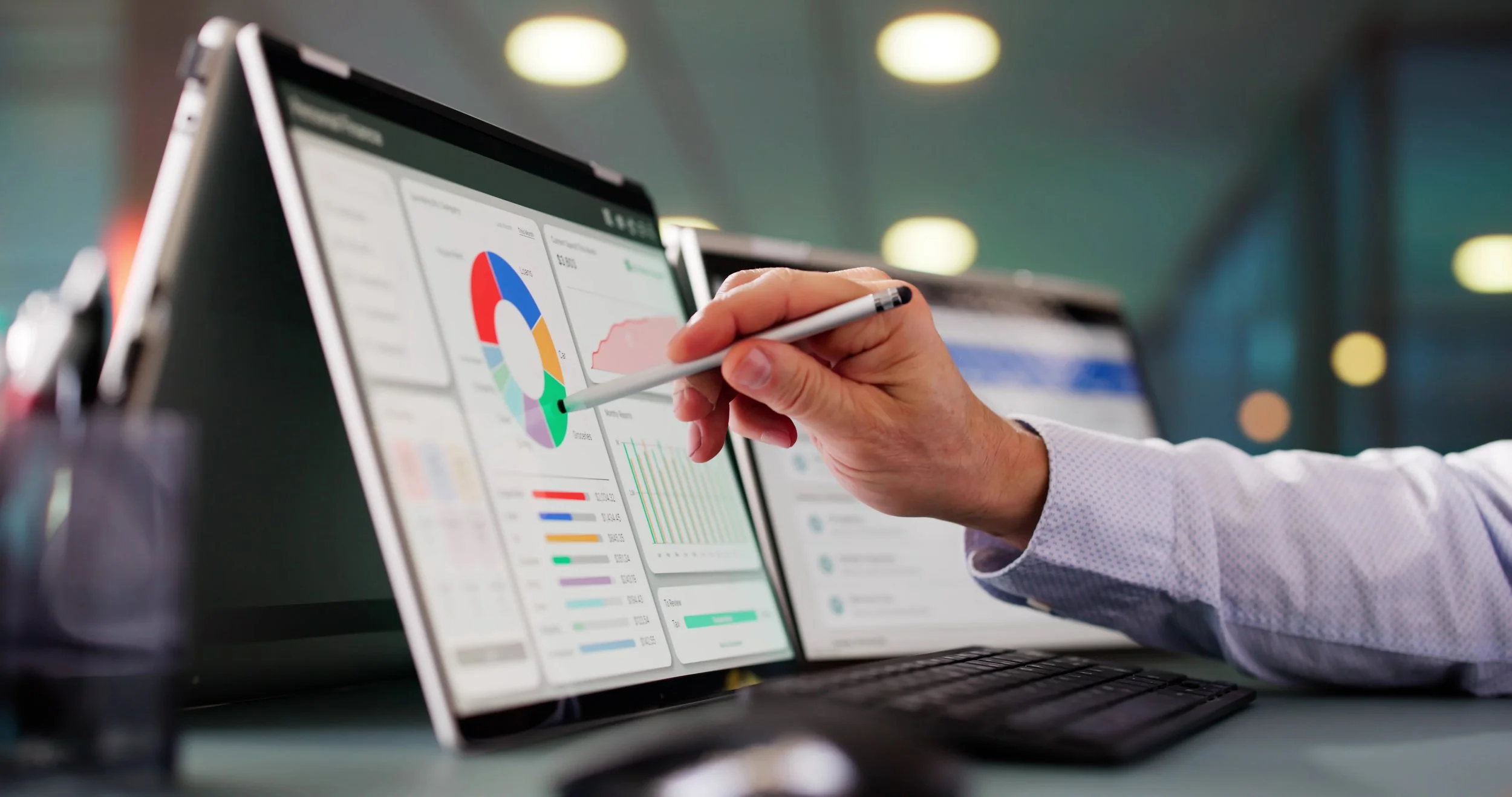 A person in a white shirt using a stylus to point at data visualizations on a dual-screen laptop, showing charts and graphs, in an office environment with blurred background lighting.