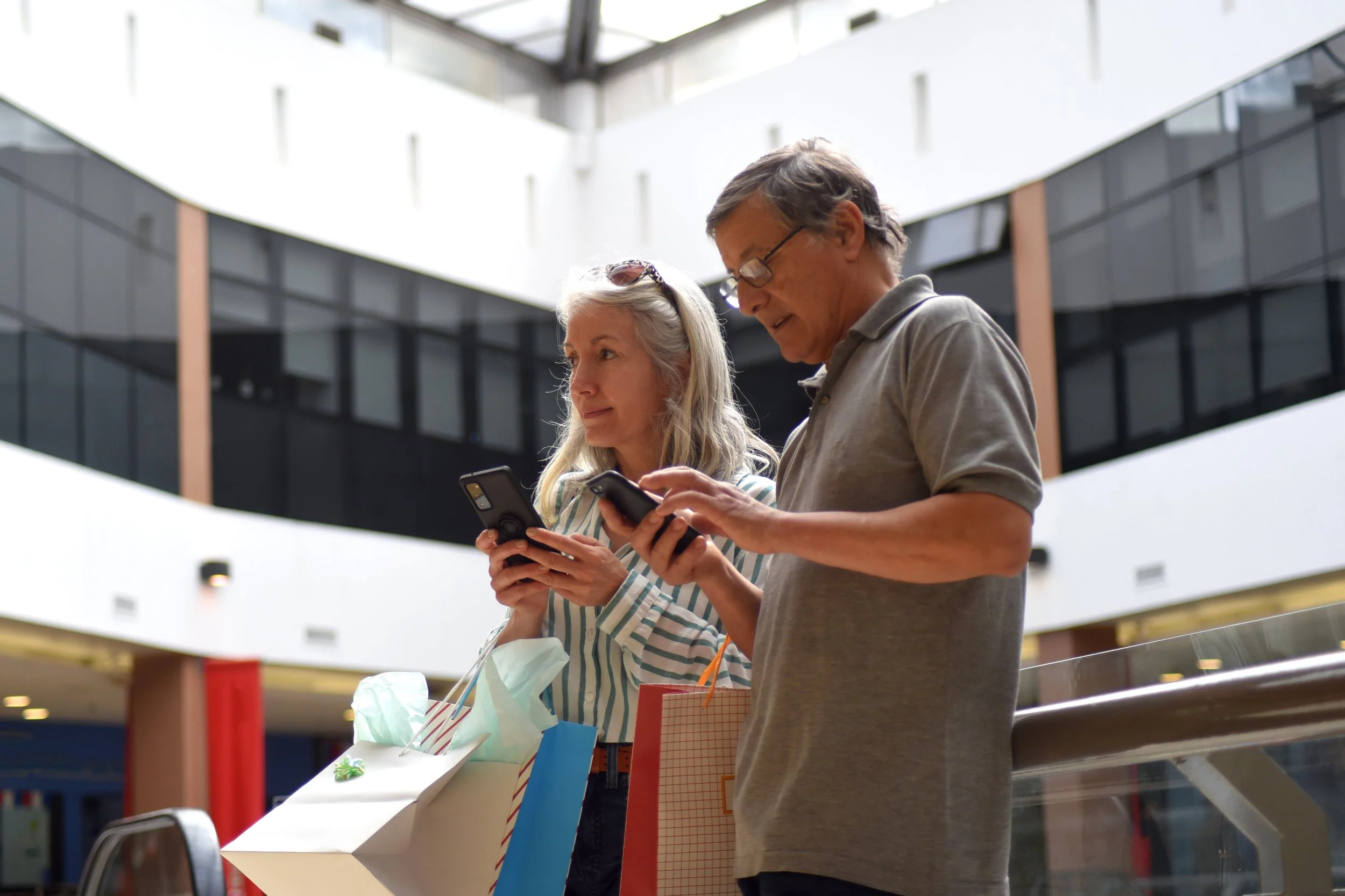 An elderly couple is standing on an escalator in a shopping mall, holding shopping bags and looking at their phones.