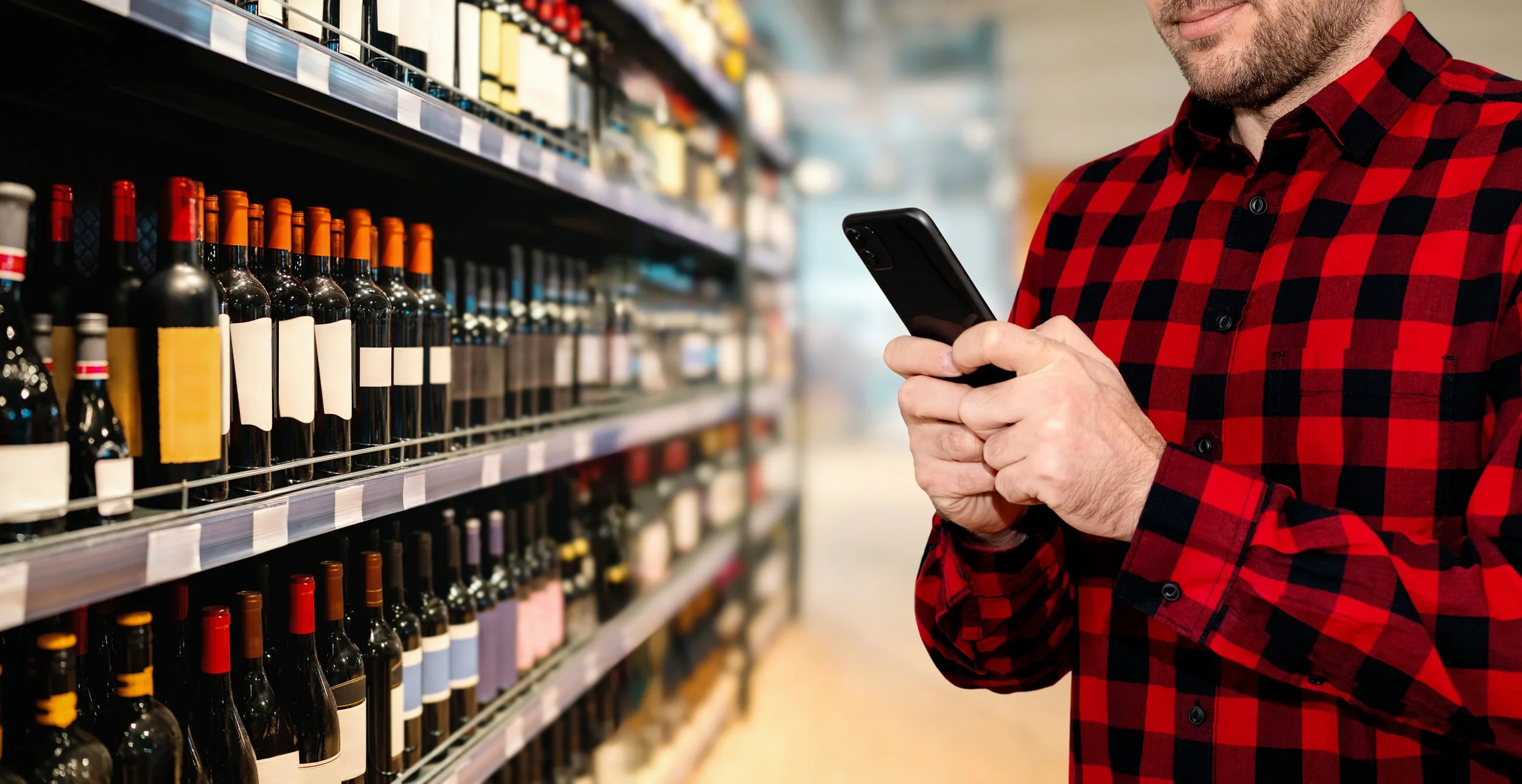 A man in a red and black checkered shirt shopping for wine at a grocery store, looking at his phone.
