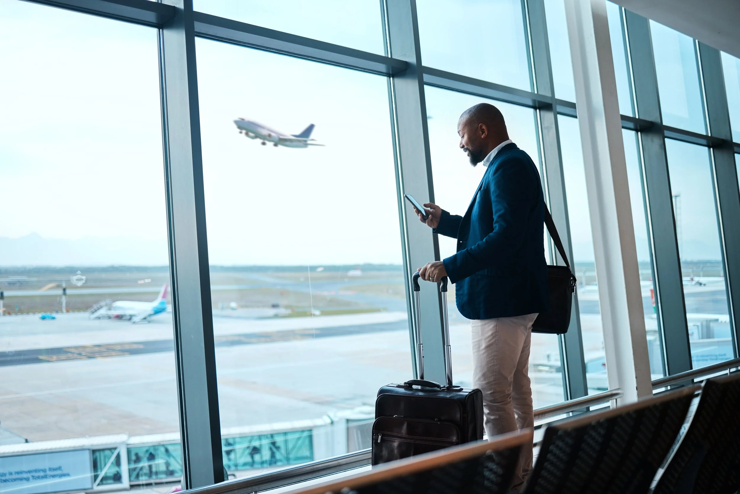 Man in a blue blazer with a suitcase at an airport, looking at his phone, with an airplane taking off outside the large glass windows.