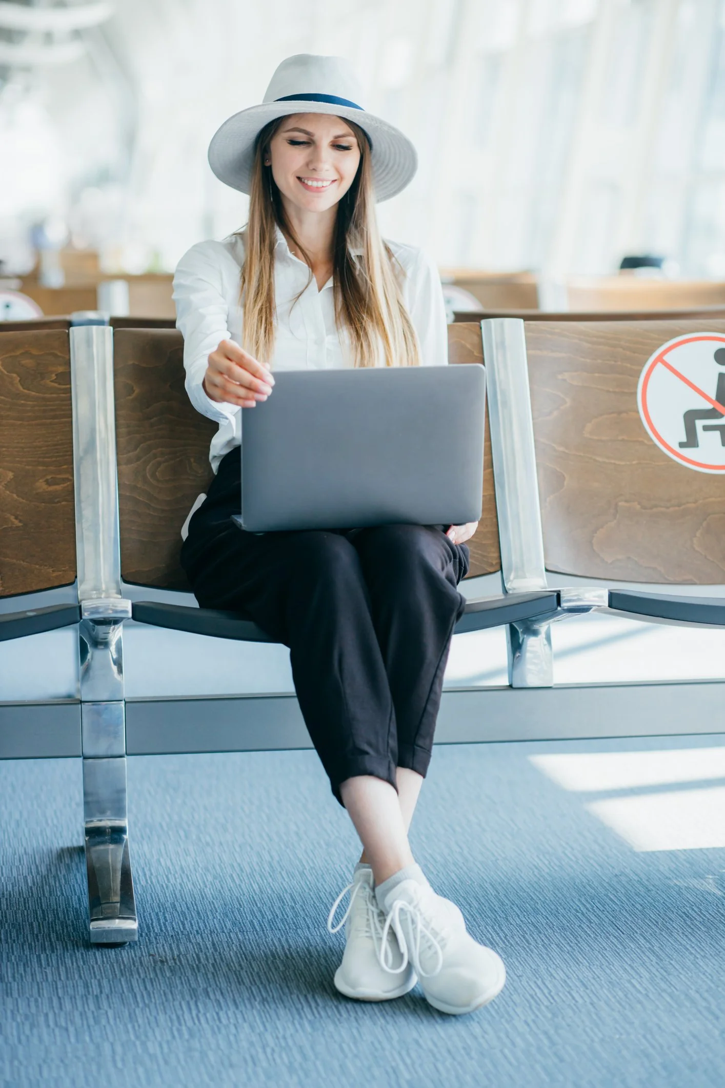A woman sitting on a bench at an airport, wearing white sneakers, a white hat, and a white shirt, working on a laptop.