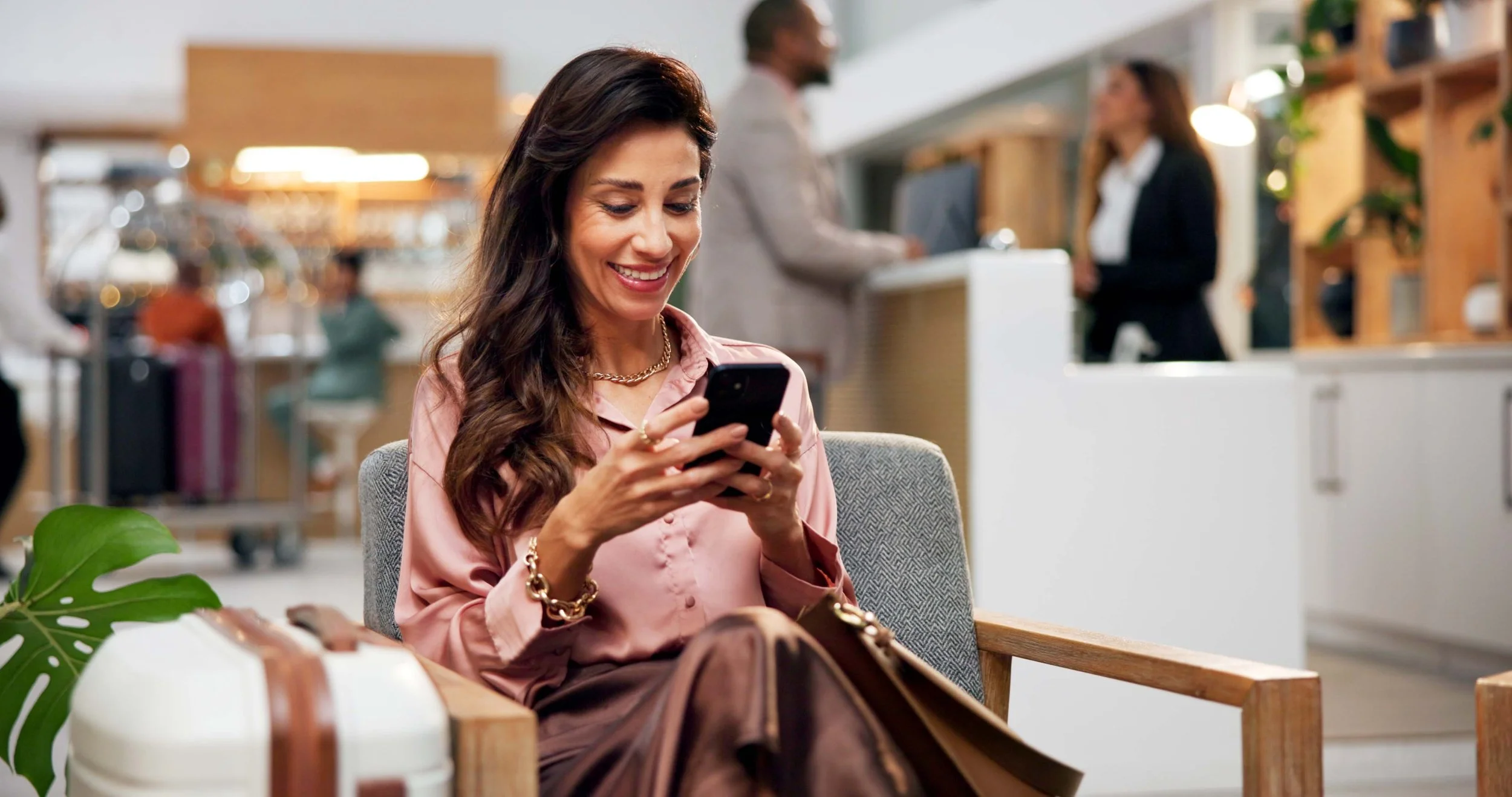 A woman sitting in a waiting area at a café or office, smiling while looking at her phone, with a busy reception area and staff in the background.