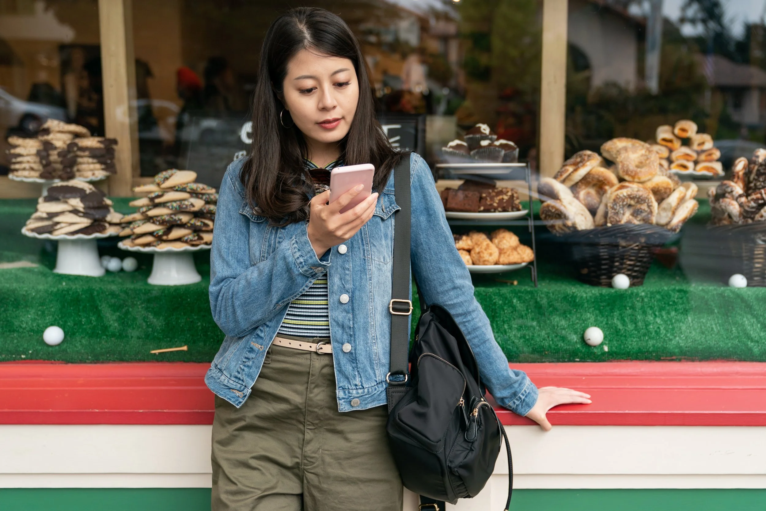 A young woman in a denim jacket and khaki pants stands outside a bakery, looking at her smartphone. Behind her are display cases filled with various baked goods and cookies.